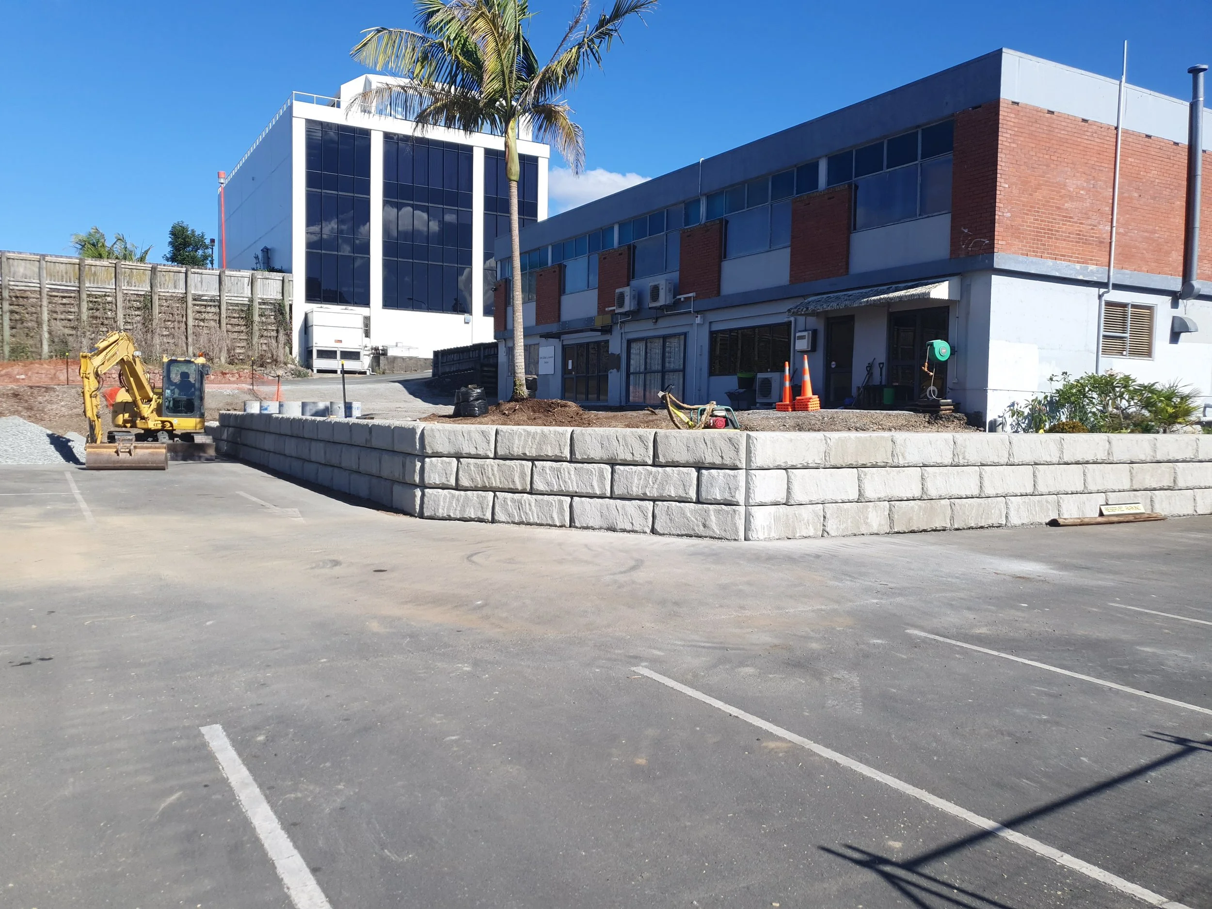 Construction site with a stone retaining wall, small excavator, and building with brick and glass exterior on a sunny day.