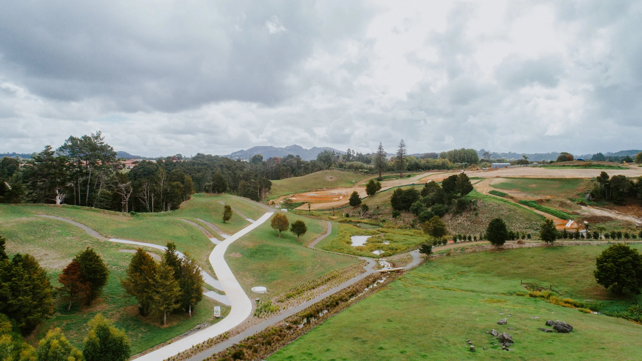Scenic landscape of rolling hills with winding paths, trees, and small ponds under a cloudy sky.