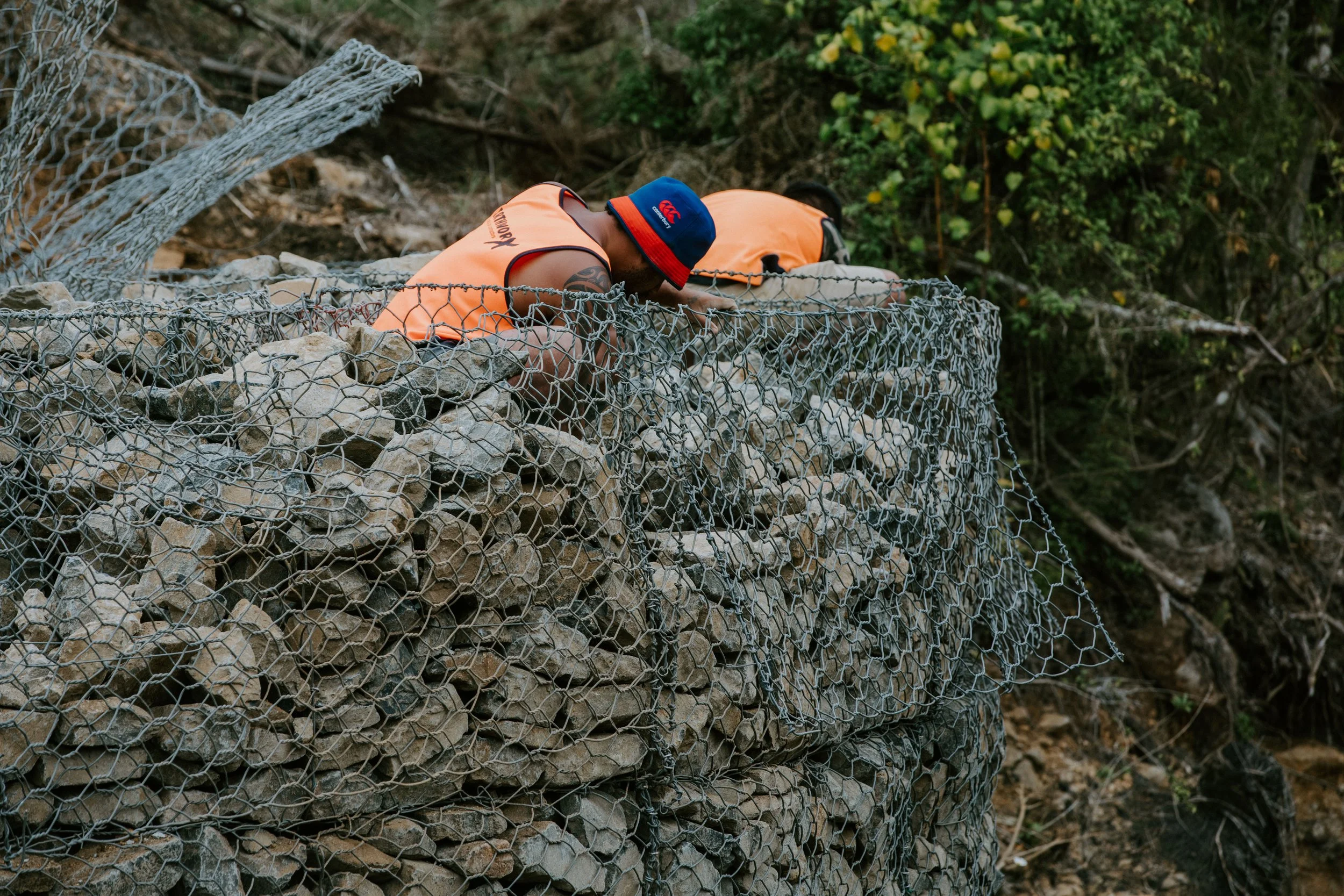 Two men wearing orange safety vests and blue hats working on a rock wall, surrounded by a wooded area.
