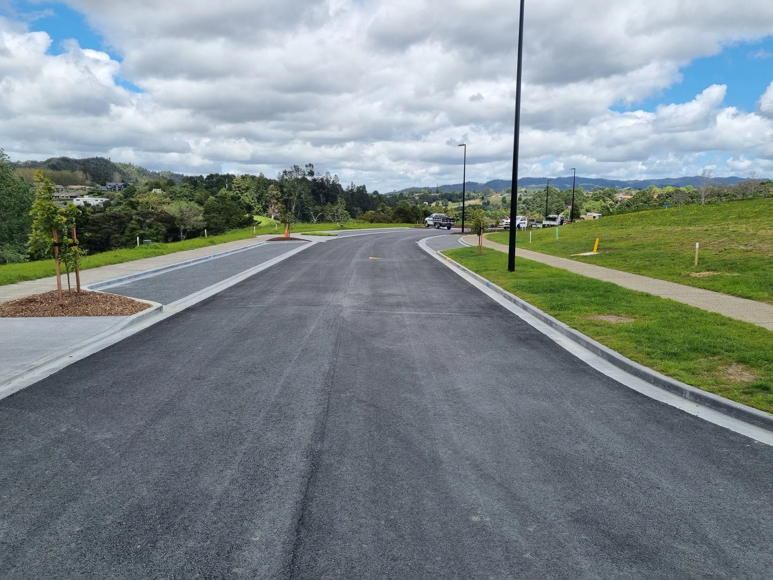 A newly paved residential street with sidewalks, streetlights, small trees in landscaped beds, green lawns, and a distant view of hills and parked cars under partly cloudy skies.