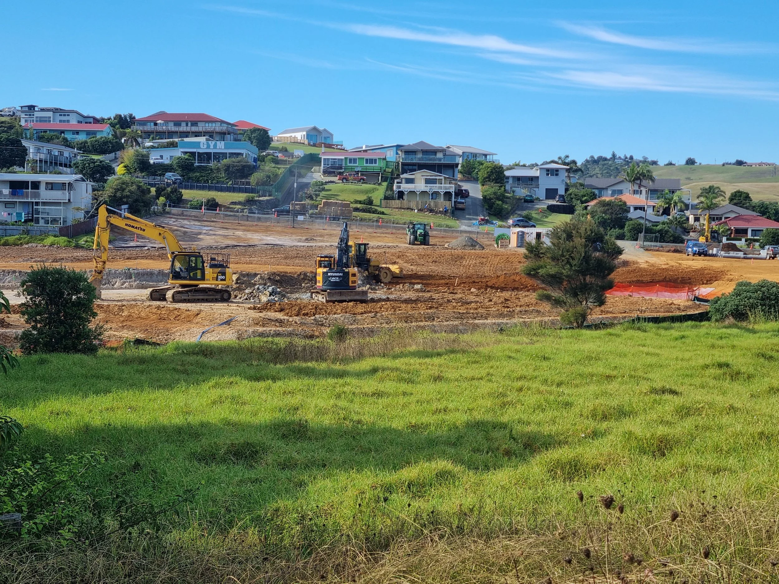 Construction site with excavators and dirt, residential houses on a hill in the background, grassy foreground, blue sky.