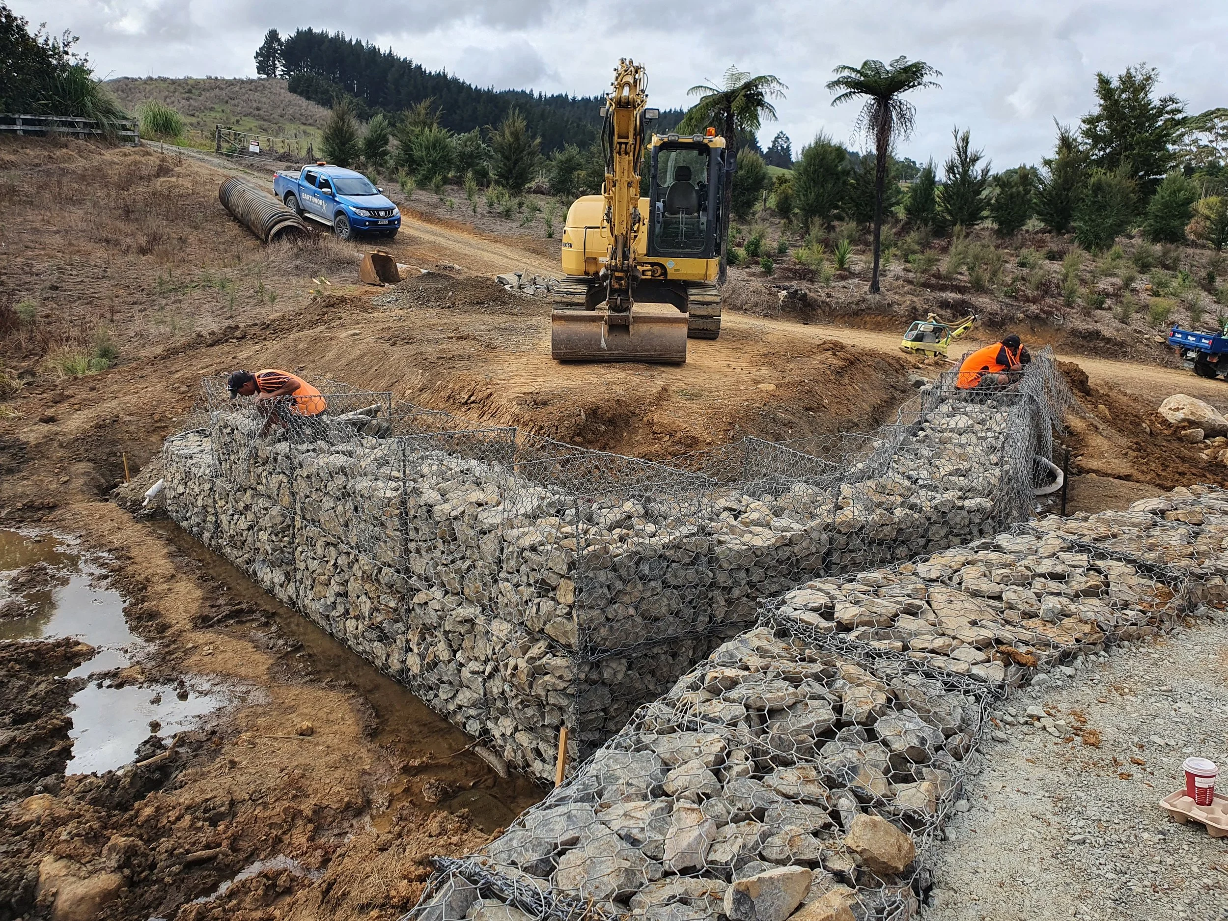 Construction workers build a riverbank reinforcement with rocks and wire mesh, a yellow excavator, and a blue pickup truck in a rural landscape.
