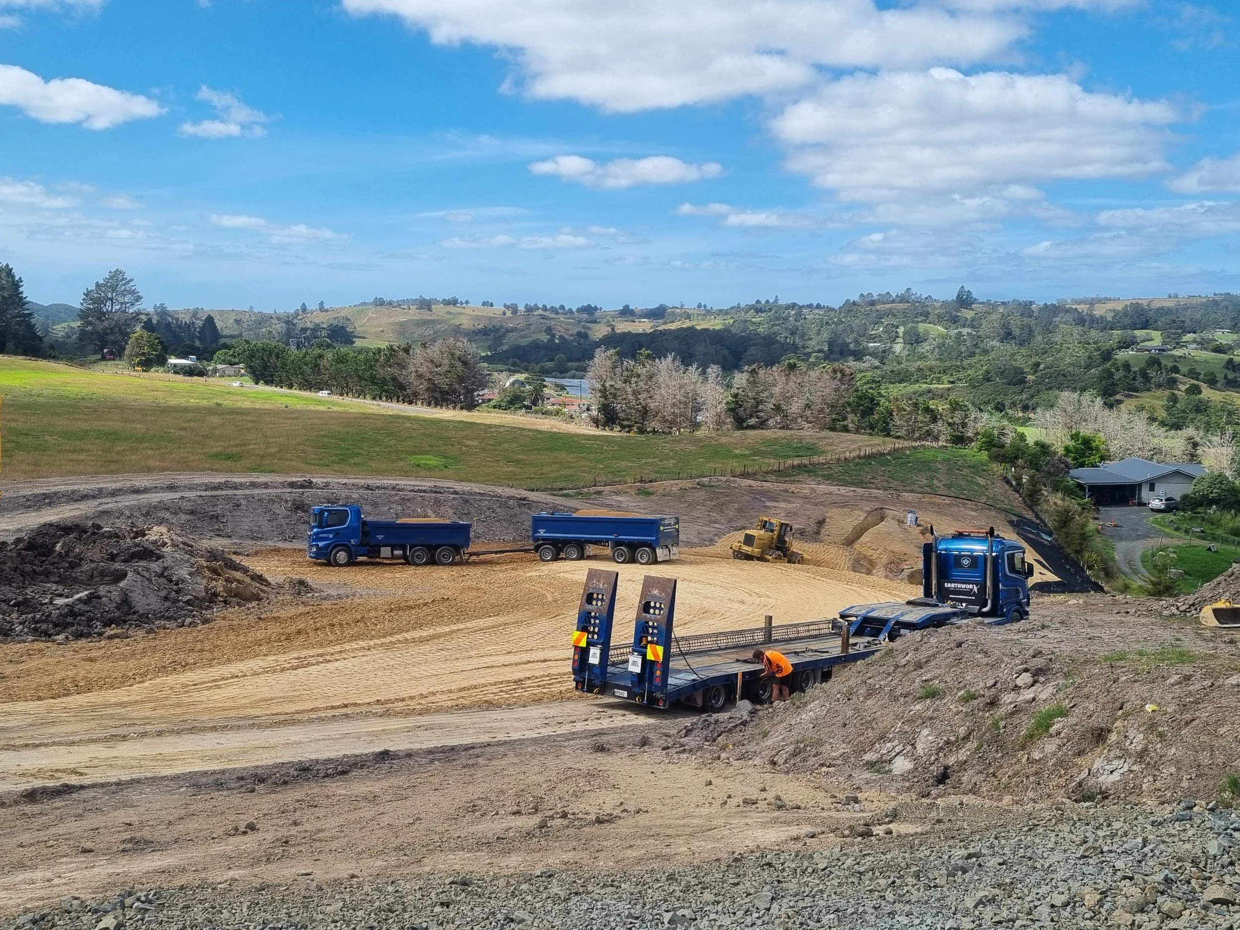 Construction site with trucks and heavy equipment on dirt in rural area with green hills and trees under a blue sky with clouds