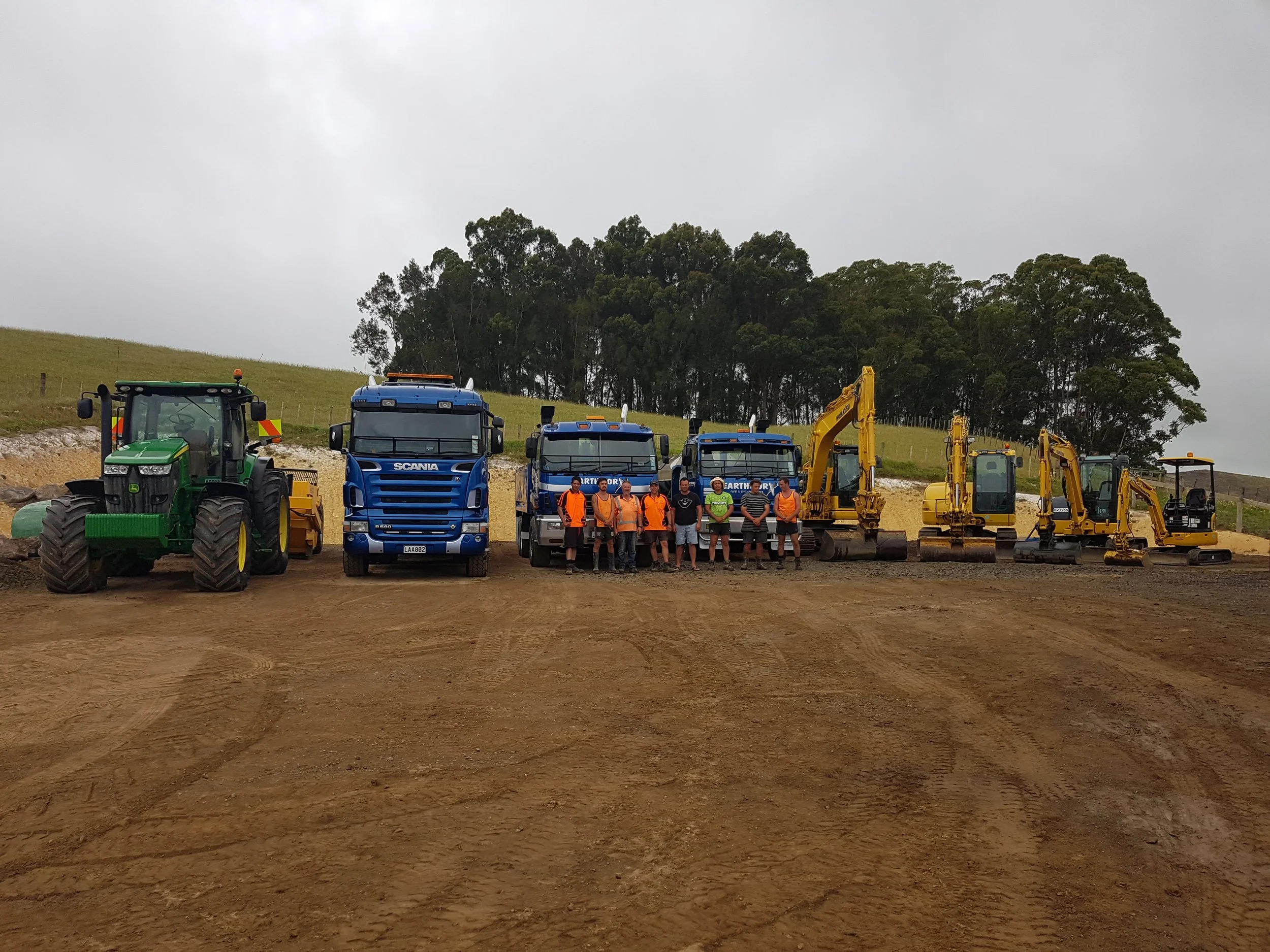 Group of construction workers and heavy machinery, including a tractor, trucks, and excavators, on a dirt construction site with trees and a cloudy sky in the background.