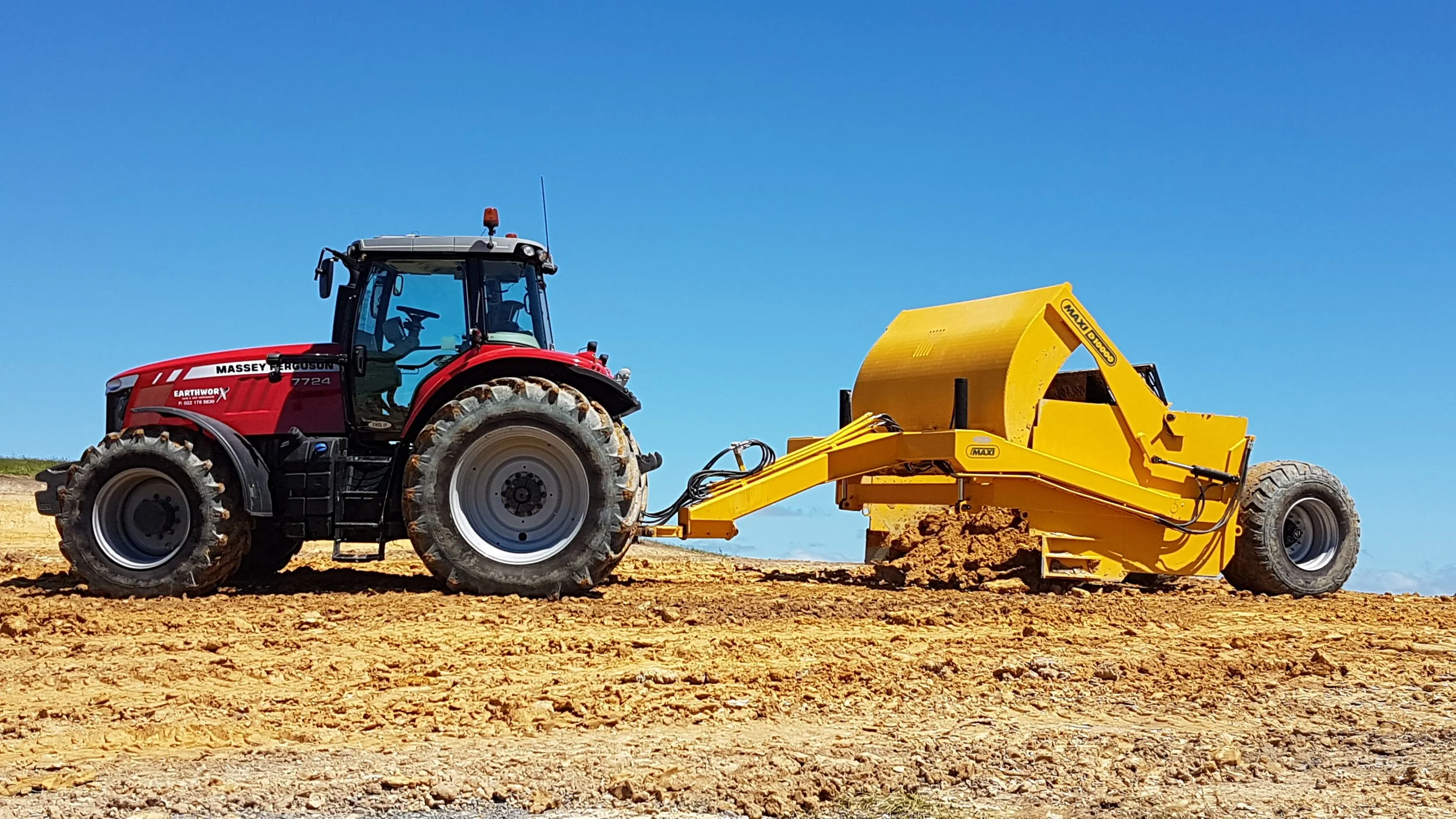 A red tractor pulling a yellow soil compactor on a construction site under a clear blue sky.
