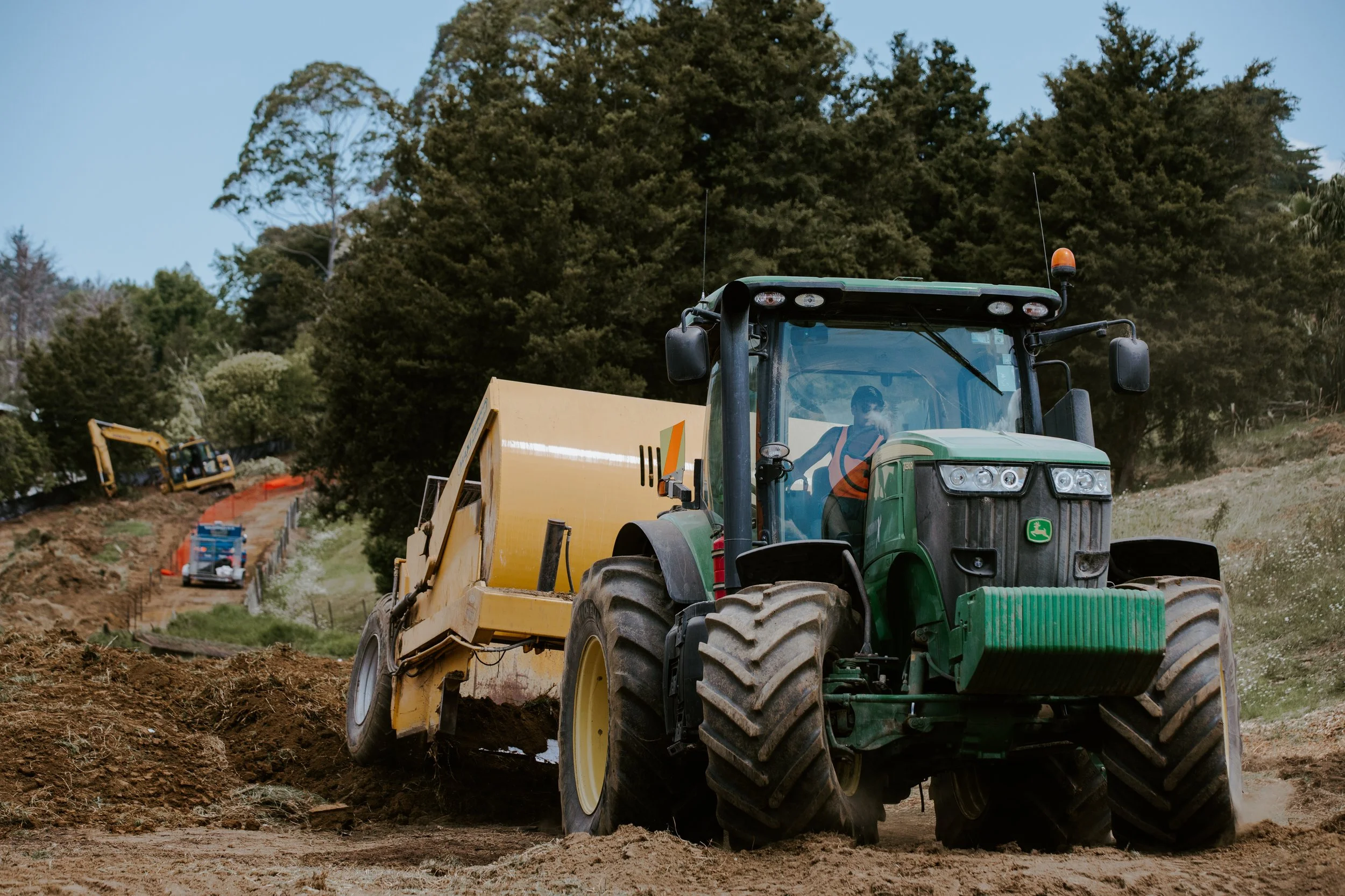 A green tractor with a yellow attachment working on a dirt construction site, with a worker inside the tractor's cab, and construction equipment in the background.