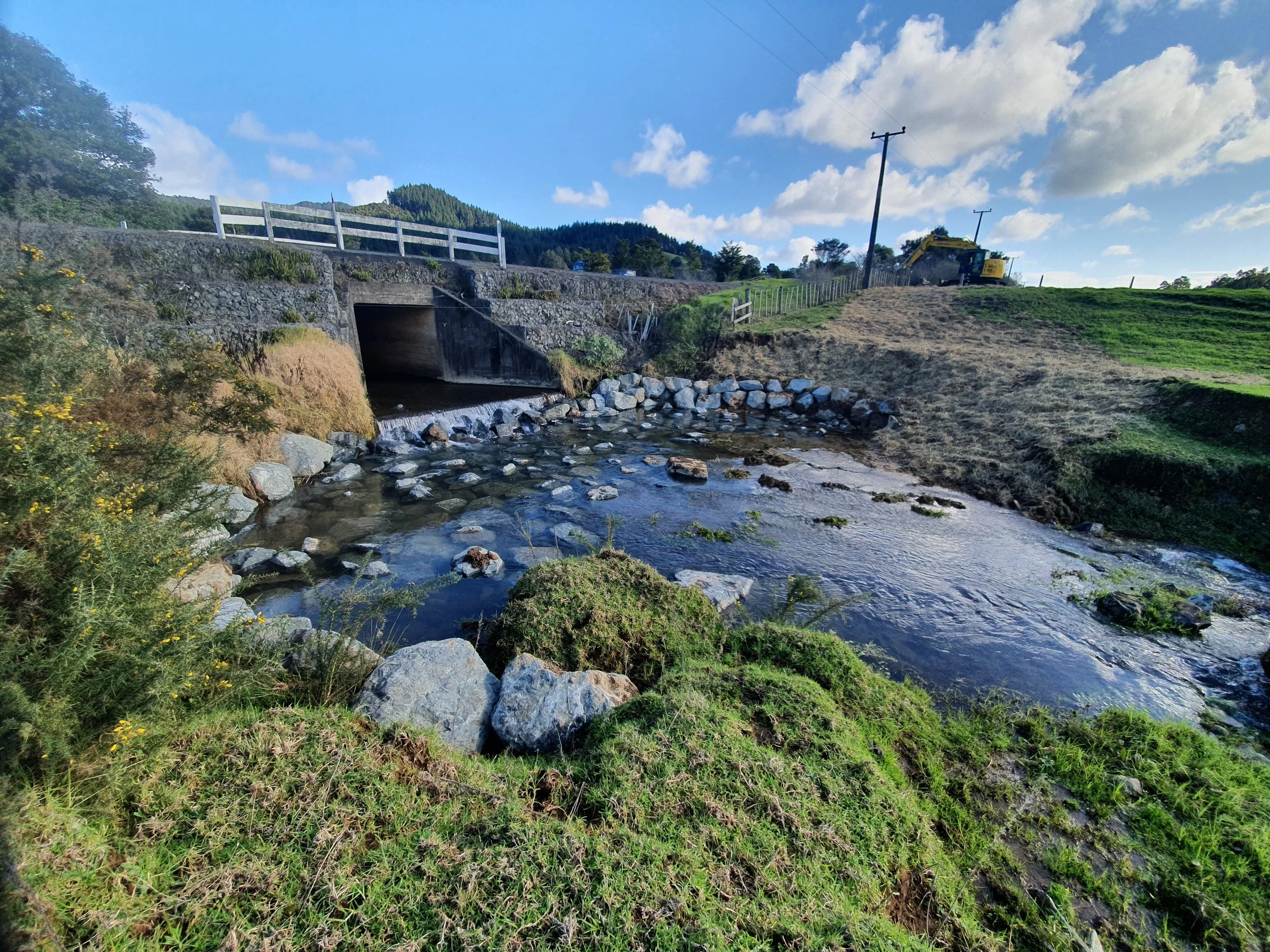 A small stream flows underneath a concrete bridge in a rural area with green grass, trees, and construction equipment nearby, under a partly cloudy sky.