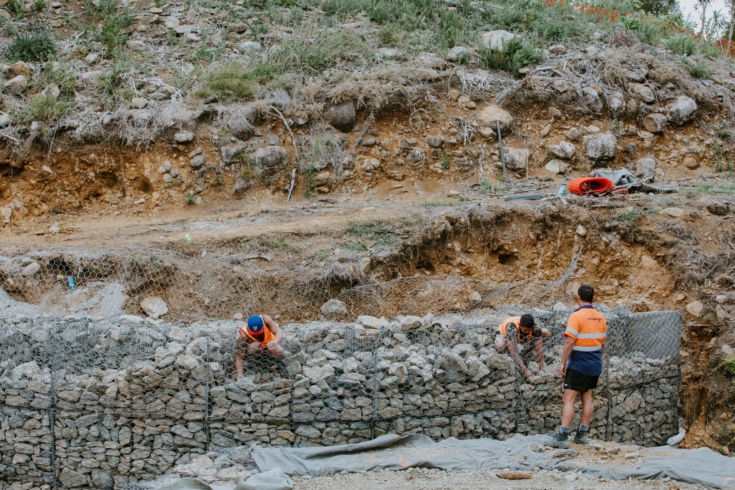 Construction workers building a rock wall with gabion baskets on a hillside.