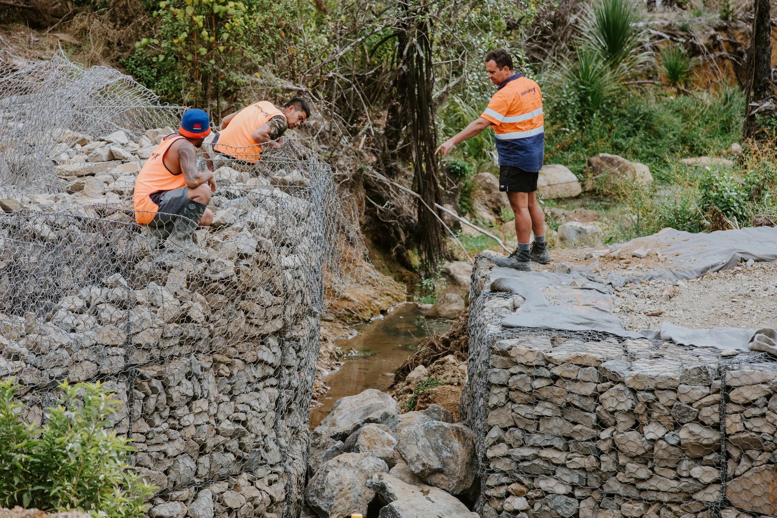 Three workers in orange vests inspecting a small creek with rocks and rocks reinforced with wire mesh and soil in a natural environment.