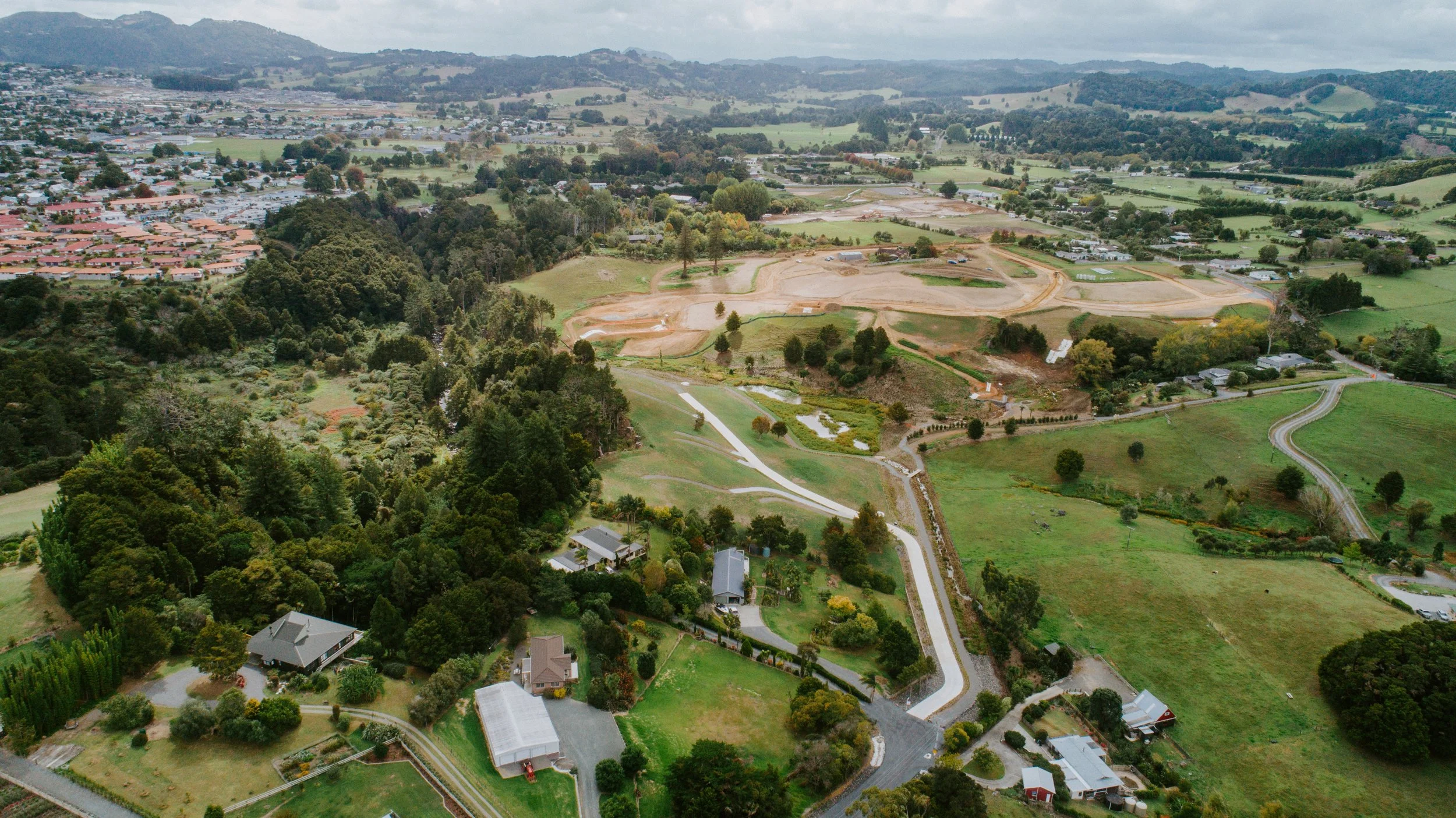 Aerial view of a rural landscape with houses, trees, winding roads, and open fields, with a city in the background and mountains on the horizon.