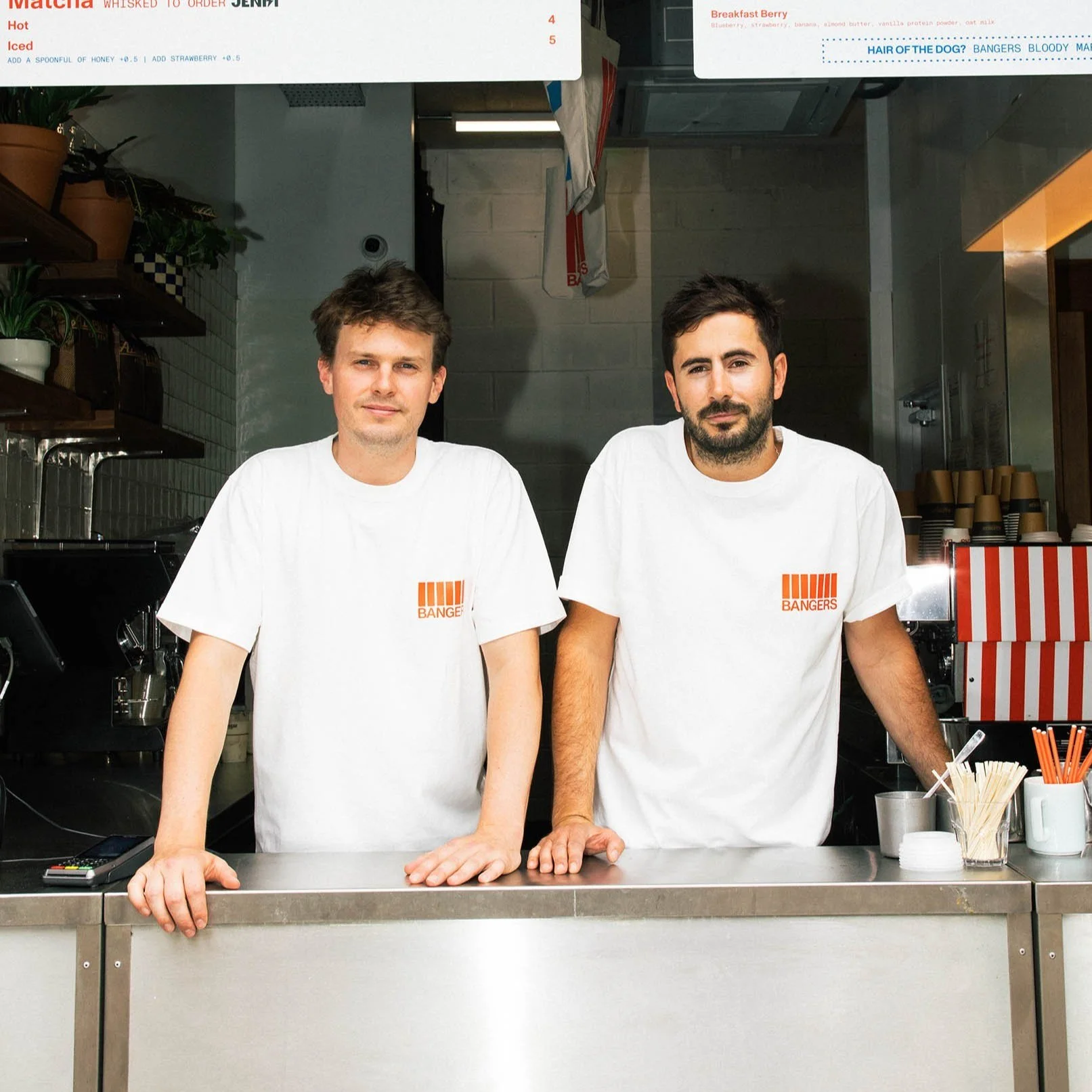 Two men standing behind a counter in a cafe or restaurant, wearing white T-shirts with an orange logo that reads 'BANGERS'.