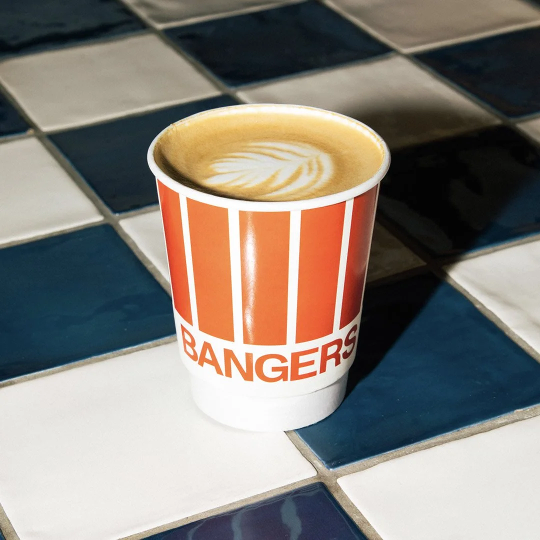 A paper cup of coffee with latte art, labeled 'BANGERS,' on a tiled table with alternating white and blue tiles.