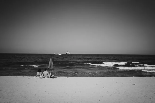 Custom Squarespace Template, Editorial Squarespace Template, Avelã Creative. Black and white photo of a couple sitting under an umbrella on a beach facing the ocean with boats in the distance.