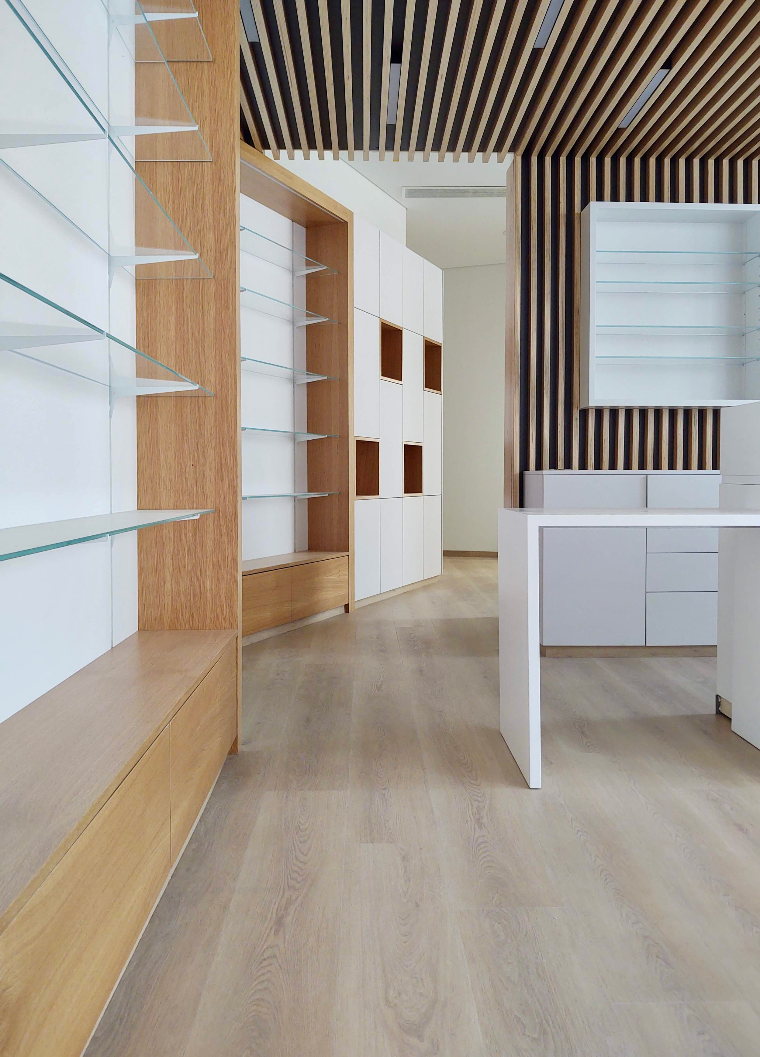 Espaço interno com paredes de alvenaria branca, detalhes de madeira clara, prateleiras de vidro, piso de madeira clara e teto com painéis de madeira escura em linhas paralelas.