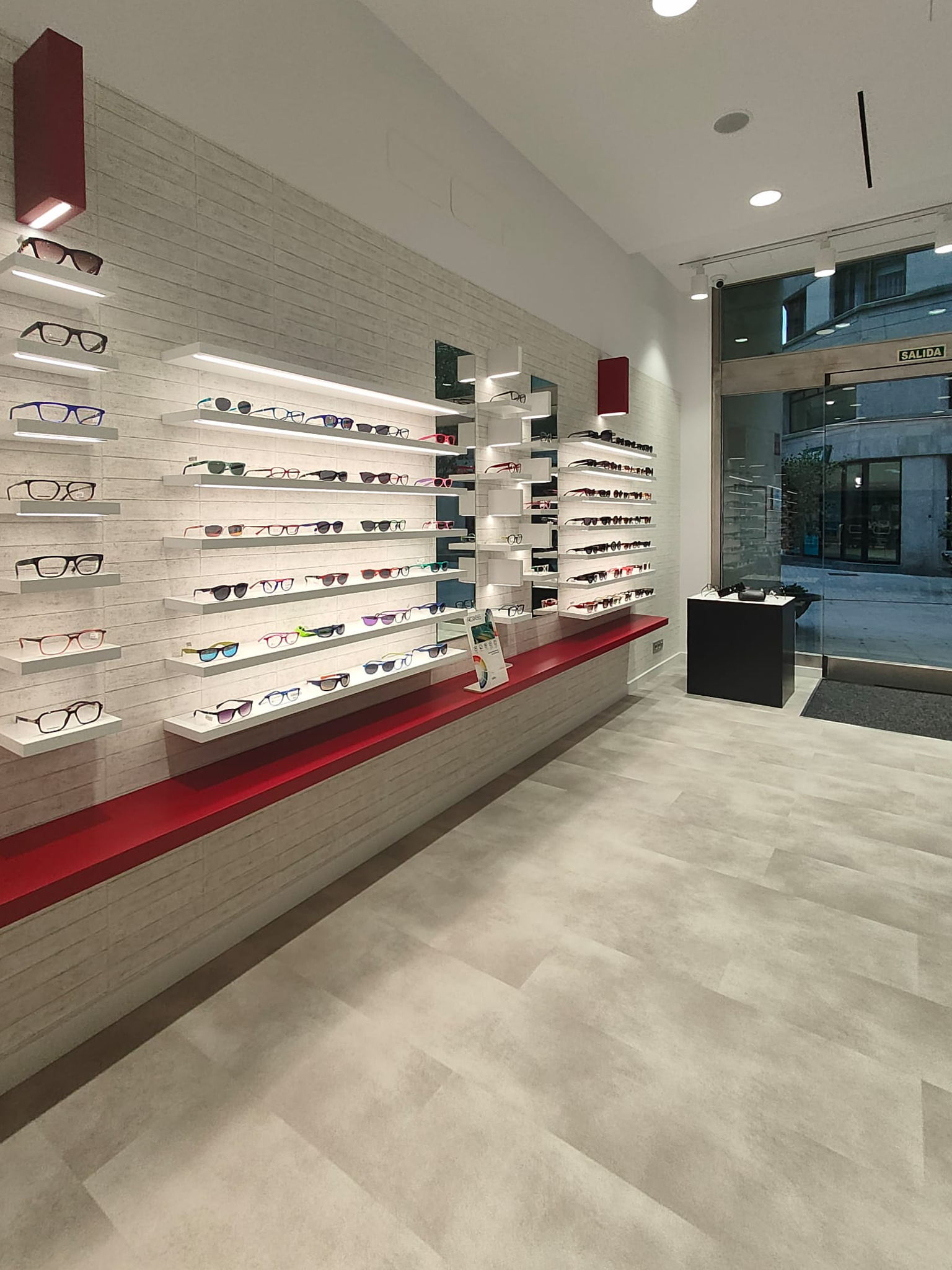 Interior de uma loja de óculos com prateleiras de óculos de diferentes estilos, cores e tamanhos, iluminação moderna e uma porta de saída de vidro na frente.