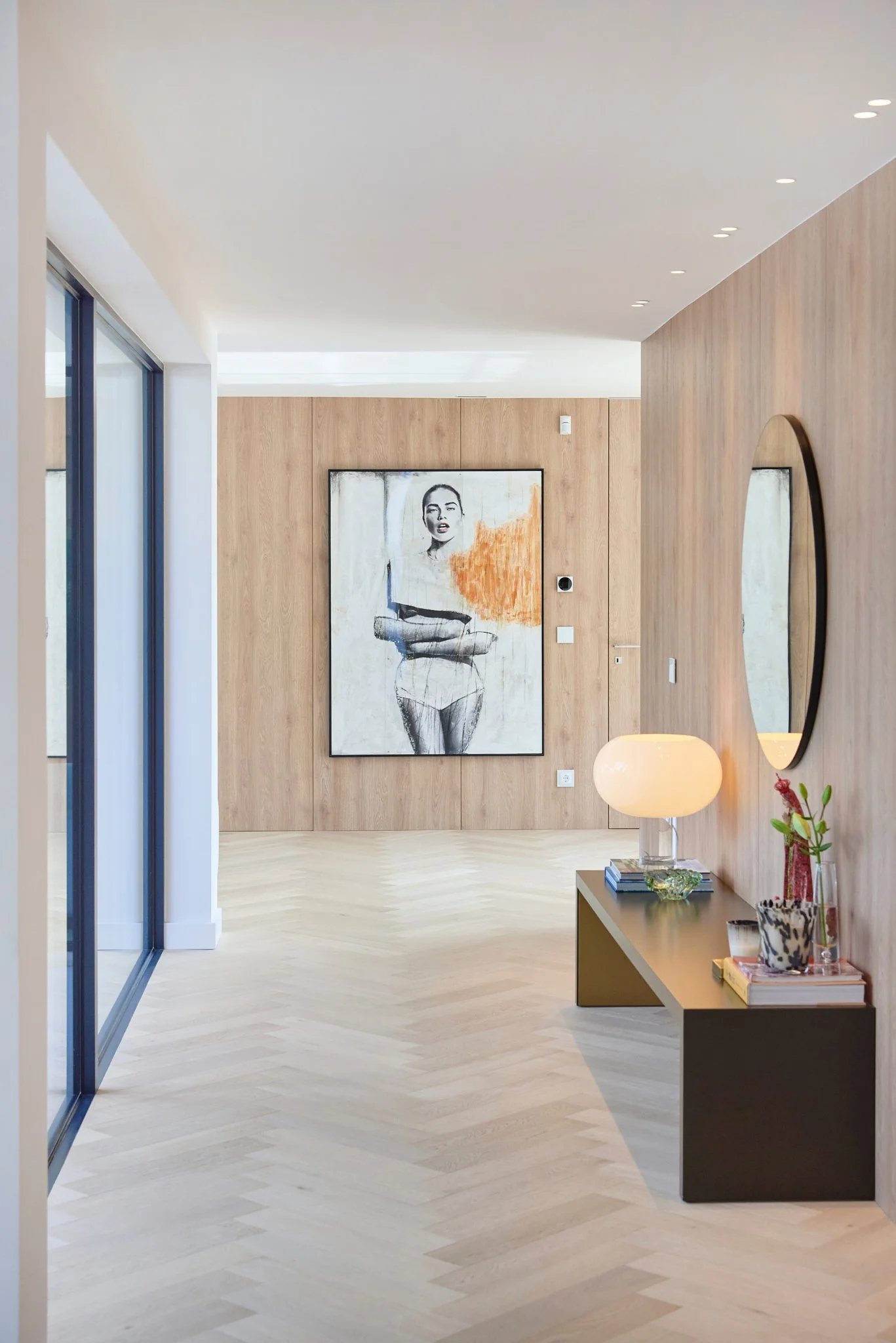 Interior de sala moderna com porta de vidro, quadro de arte na parede e mesa decorativa com lâmpada, livros e vasos de flores.