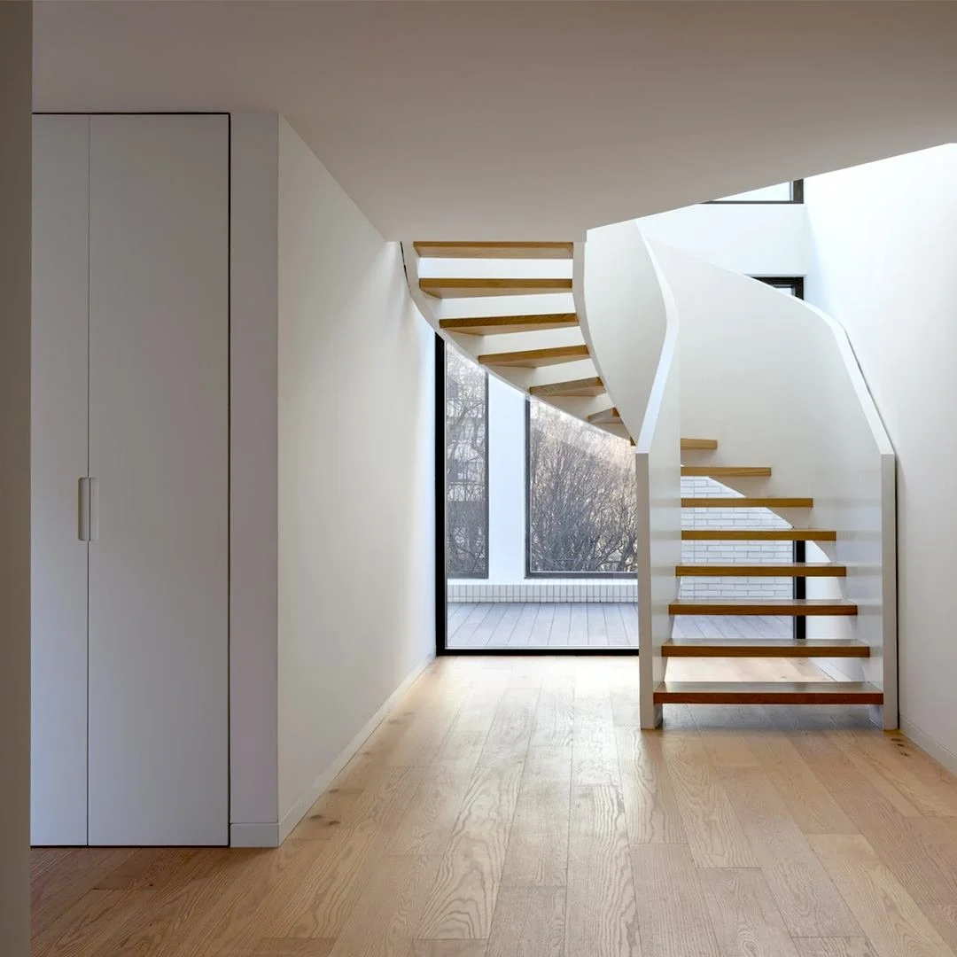 Escada de madeira branca e lâminas de madeira no piso de uma casa com janelas grandes de vidro, parede branca e varanda ao fundo.