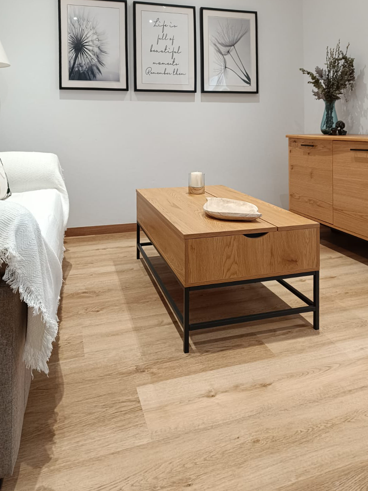 Sala de estar decorada com quadro na parede, mesa de centro de madeira com gaveta, sofá com manta branca, e armário de madeira com vaso de flores ao lado.