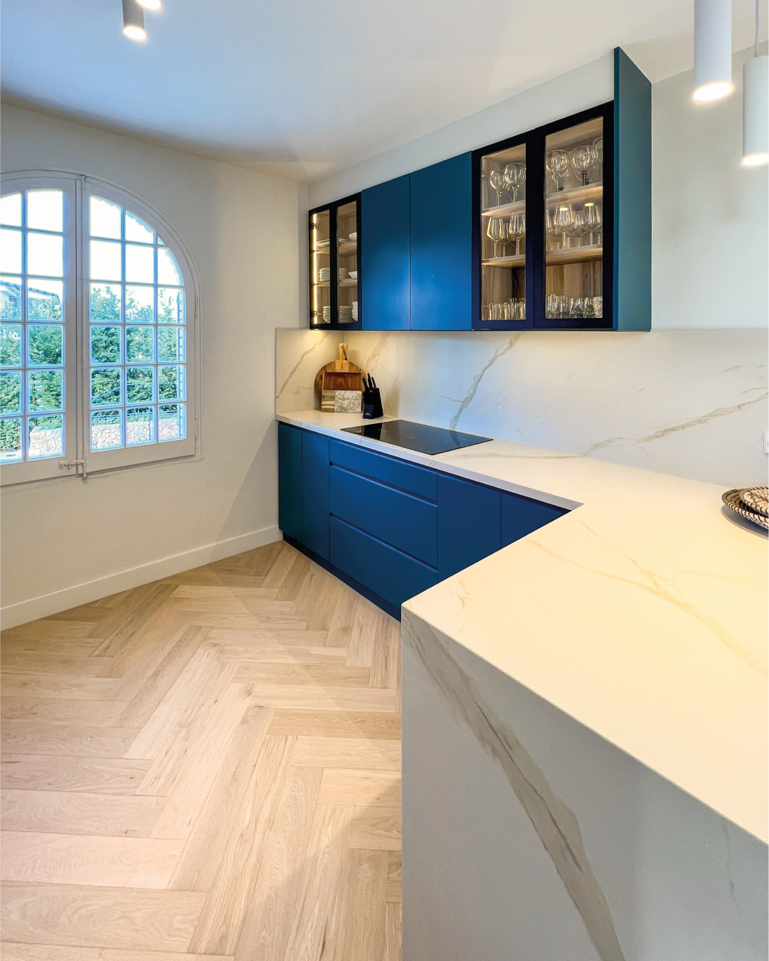 Cozinha moderna com móveis azuis, armários superiores de vidro e uma bancada de mármore, janela de ferro com vista para árvores verdes.