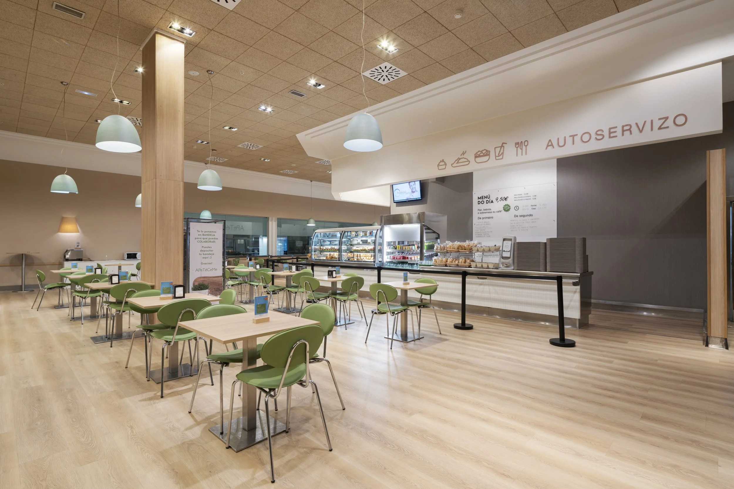 Salão de restaurante com mesas e cadeiras verdes, balcão de autoatendimento com alimentos e um painel de menu, iluminação moderna e piso de madeira.