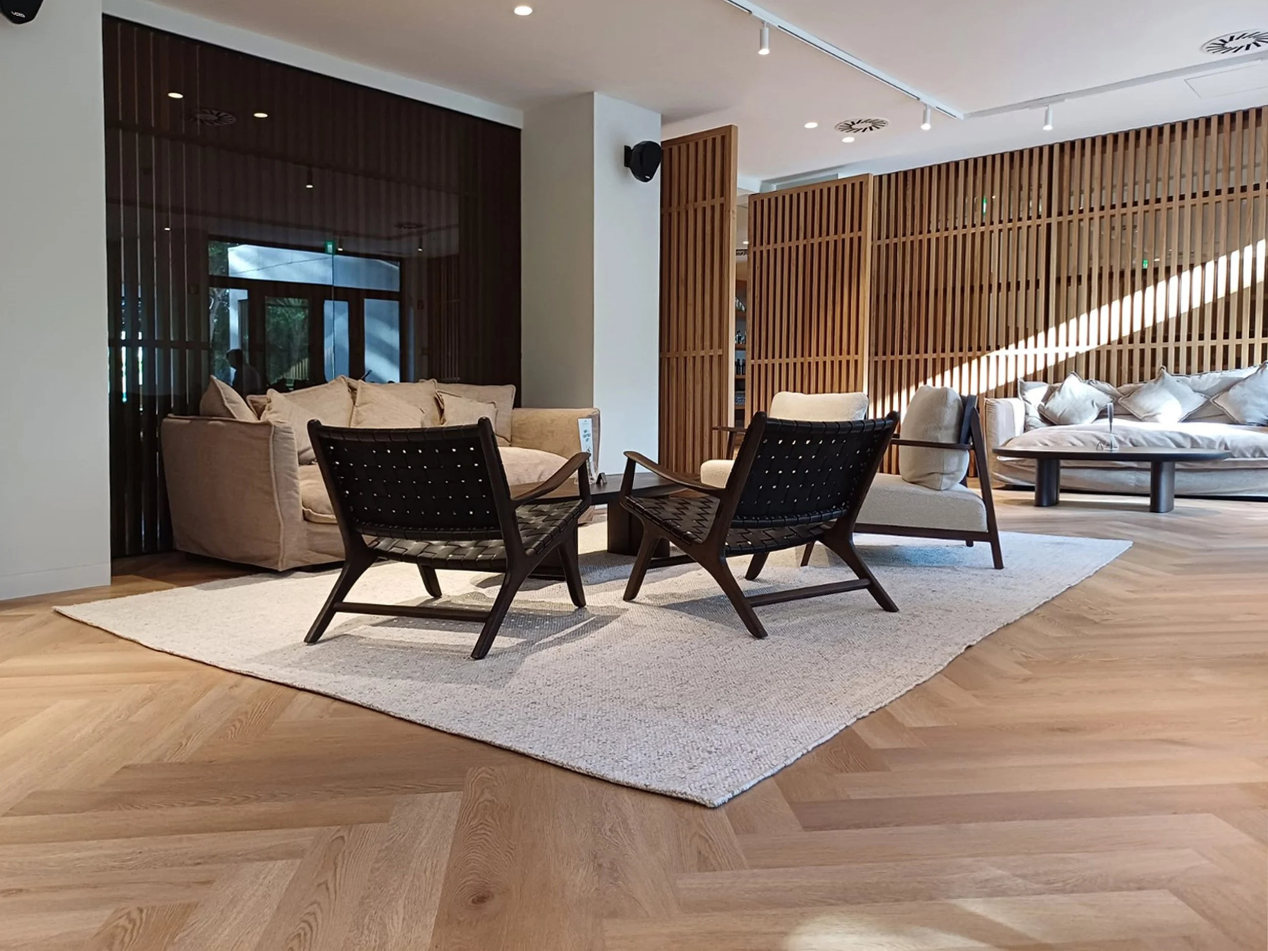 Sala de estar moderna com móveis de madeira clara, sofá bege, cadeiras pretas com estrutura de madeira, tapete claro, decoração minimalista e iluminação natural com raios de sol.