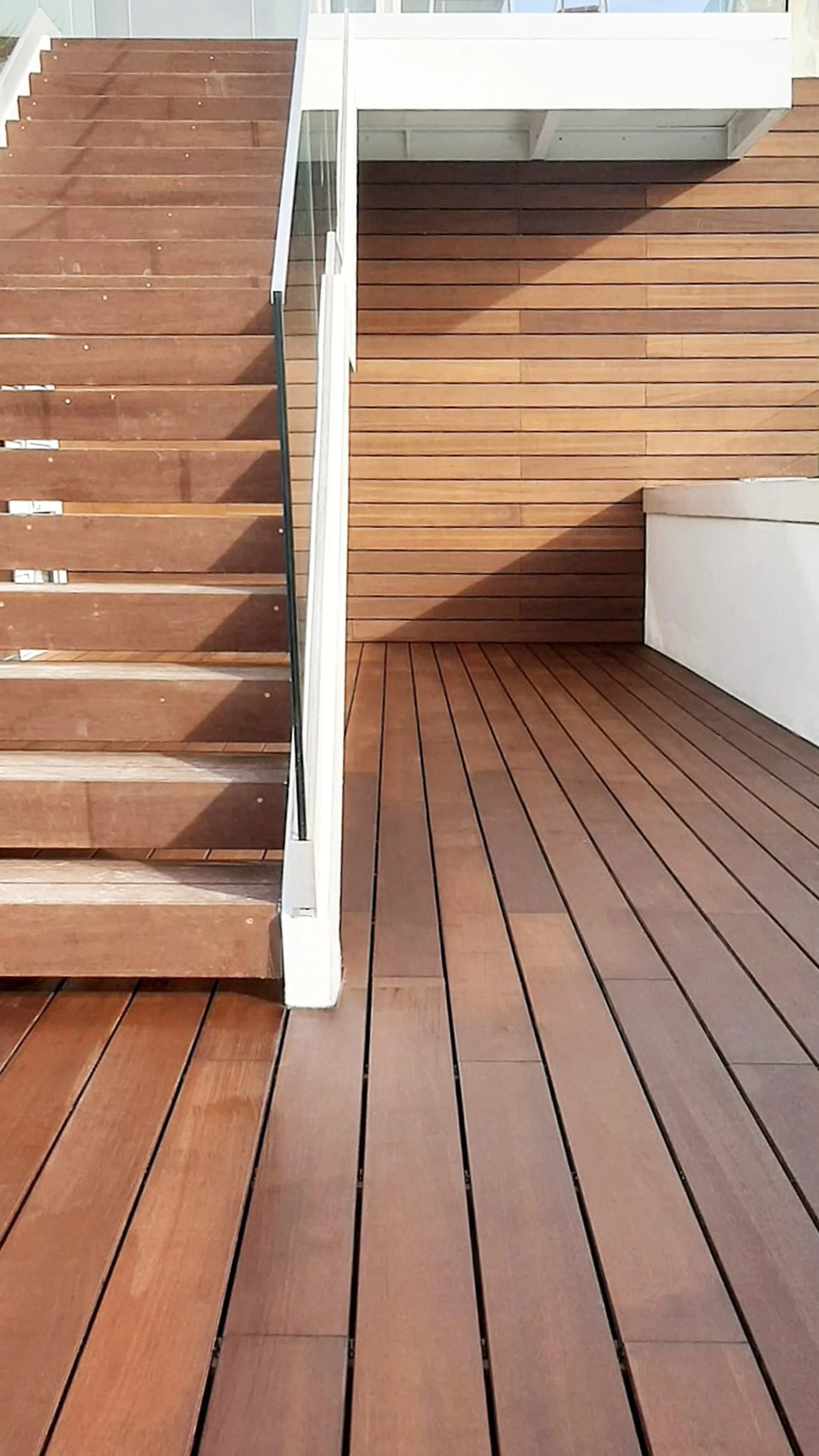 Escada de madeira com degraus e corrimão branco ao lado, e área de piso de madeira no chão ao redor, parede de madeira ao fundo.