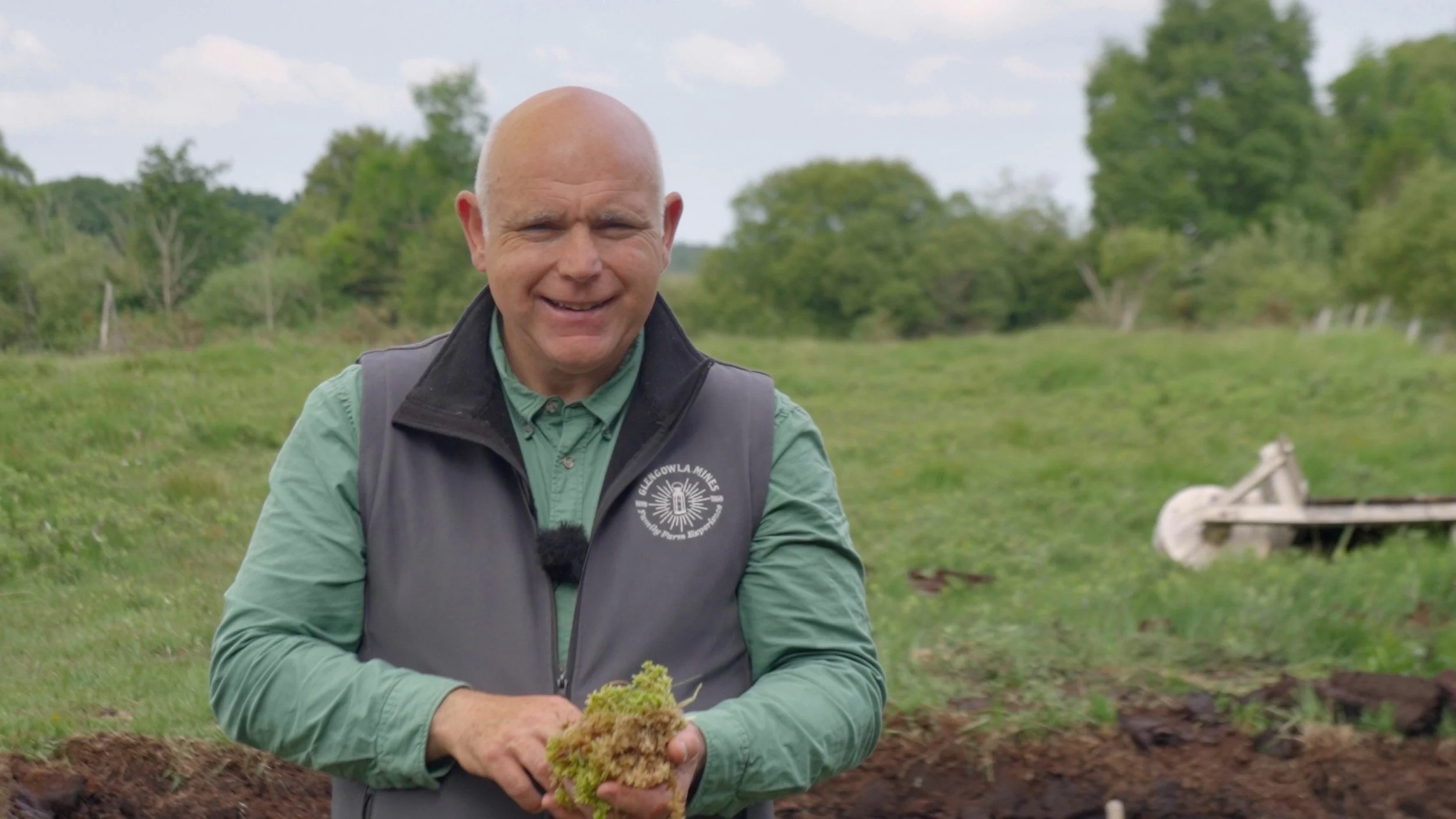 A man outdoors in a grassy field holding moss and soil, smiling at the camera, with trees and cloudy sky in the background.