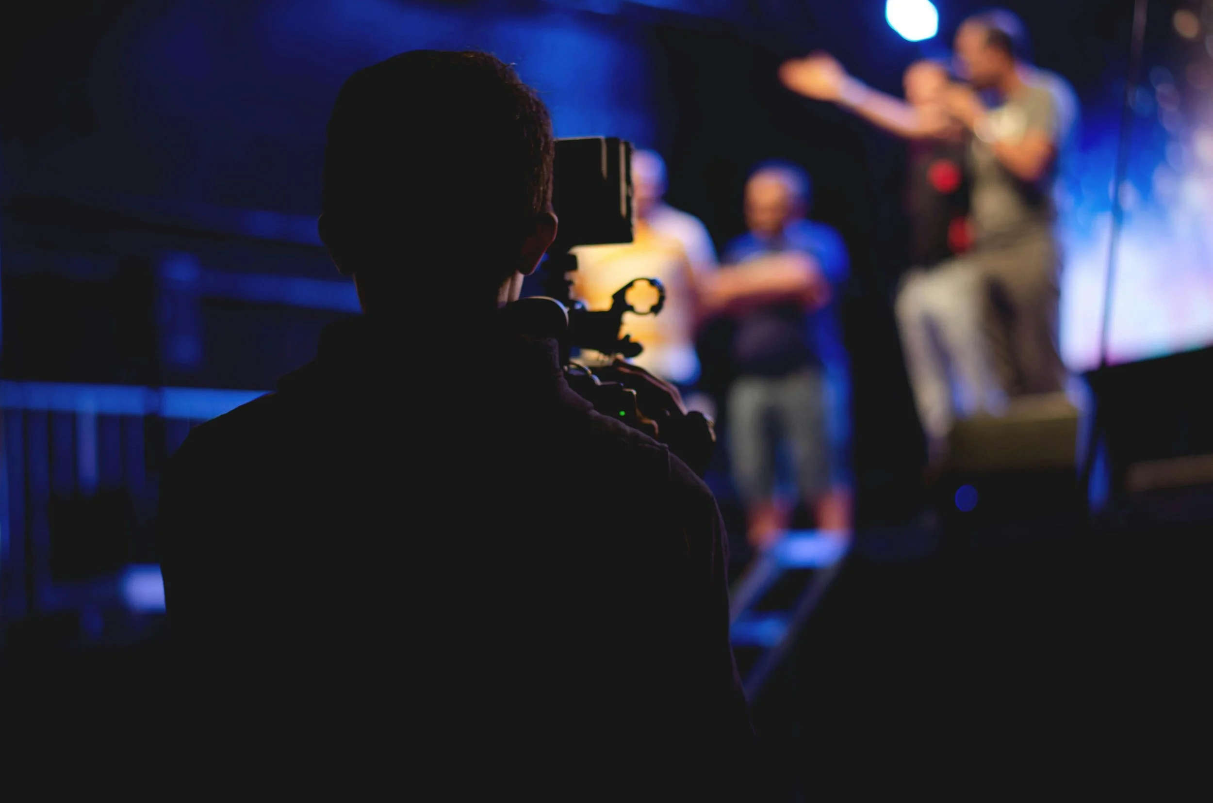 A person filming a stage performance with a camera, silhouettes in the foreground, colorful stage lights, and performers on stage in the background.