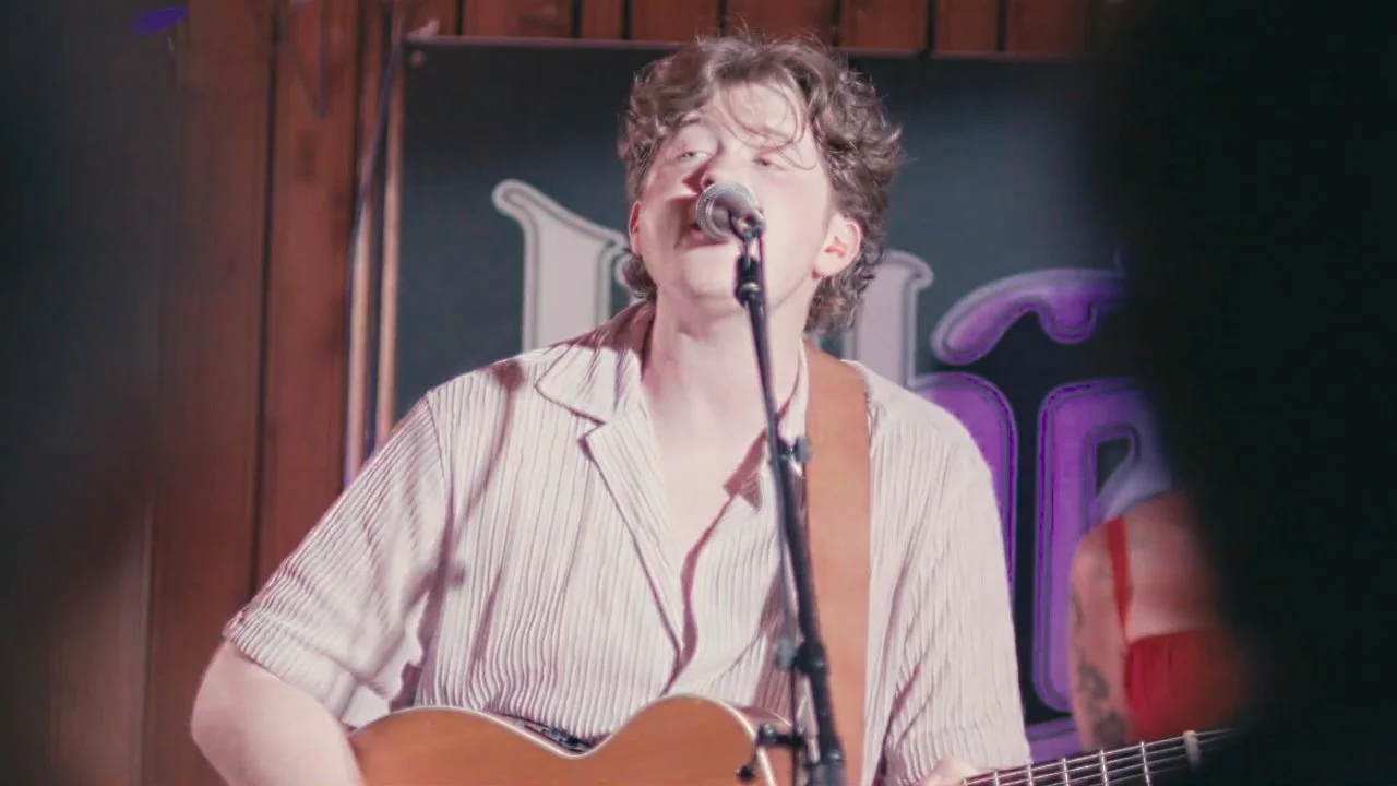 A young man with curly hair singing into a microphone while playing an acoustic guitar in a dimly lit venue.