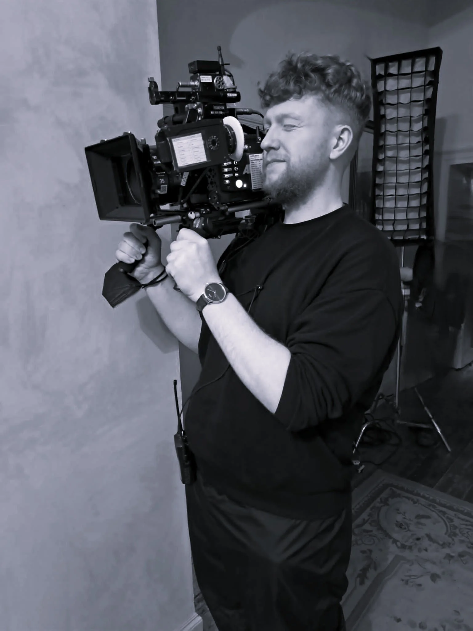 A man operating a professional video camera indoors, wearing a black shirt and a watch, with filming equipment and lighting in the background.