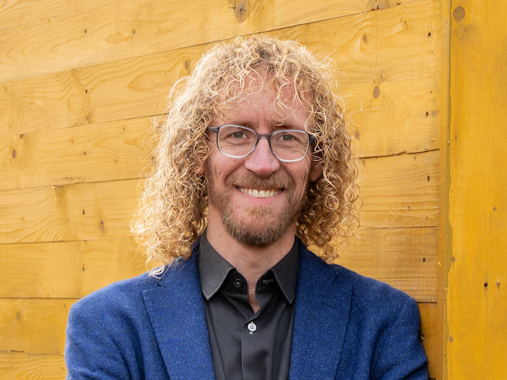 Portret van een lachende man met grijs haar, gekleed in een grijs jasje, voor een groene achtergrond met water en bomen.