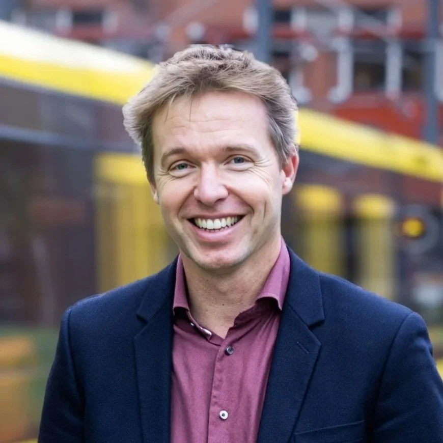 Een lachende man met lichtbruin haar en een rood overhemd met bloemenpatroon, voor een achtergrond met grote ramen.