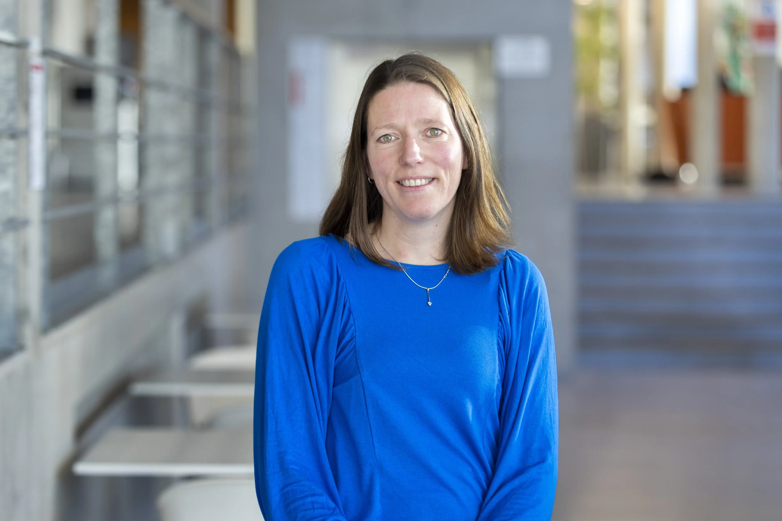 Portret van een vrouw met bruin haar in een blauw overhemd, lachend, in een moderne, open kantooromgeving.
