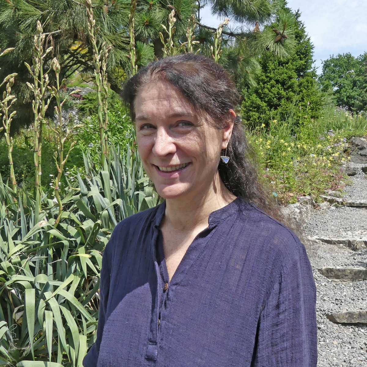 Portret van een vrouw met bruin haar en zwarte ete die in een park of bos staat, smiling, met een groene achtergrond.