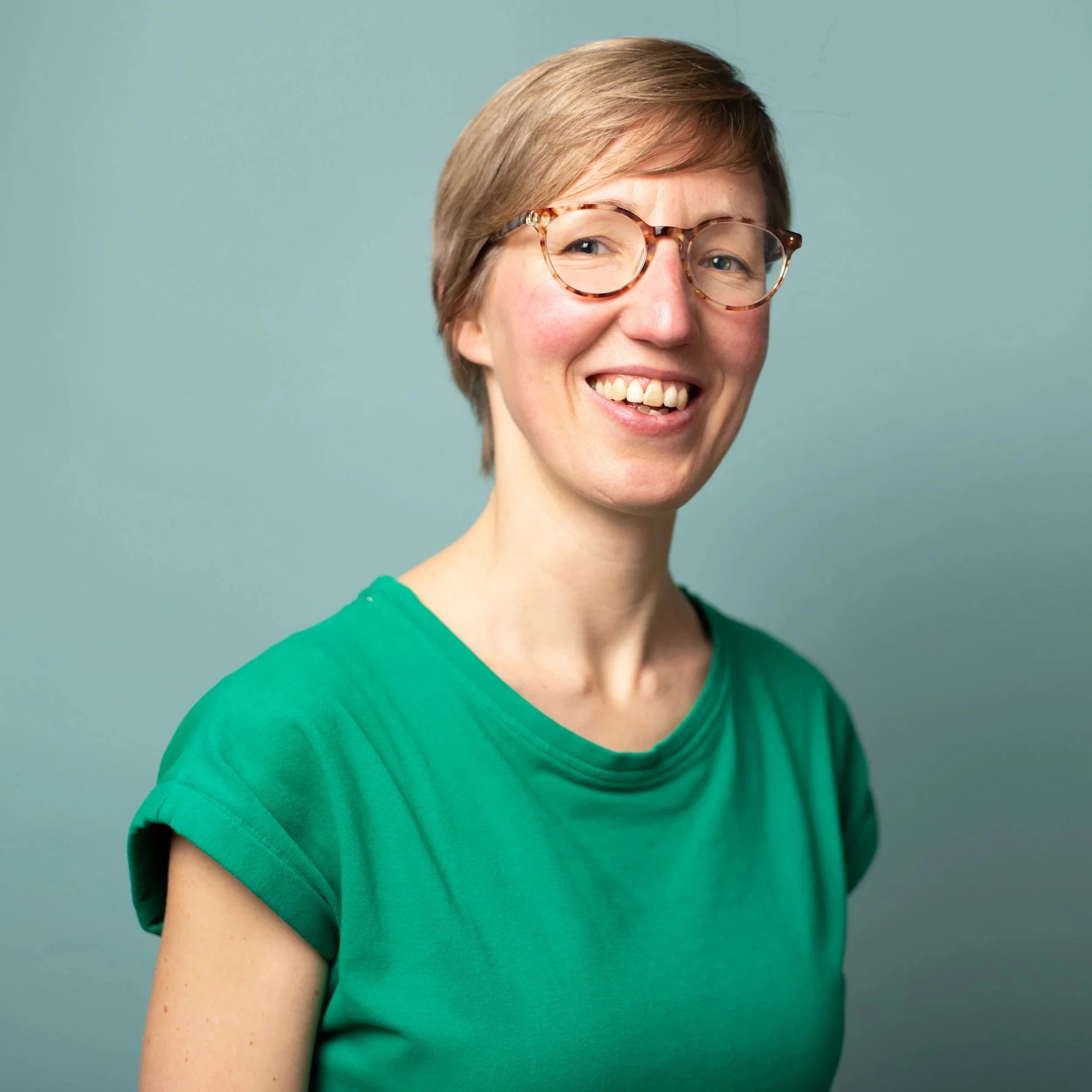 Portret van een lachende vrouw met korte bruin blonde haar, bril met een tortoise rand, en een groene T-shirt, voor een effen lichtgroene achtergrond.