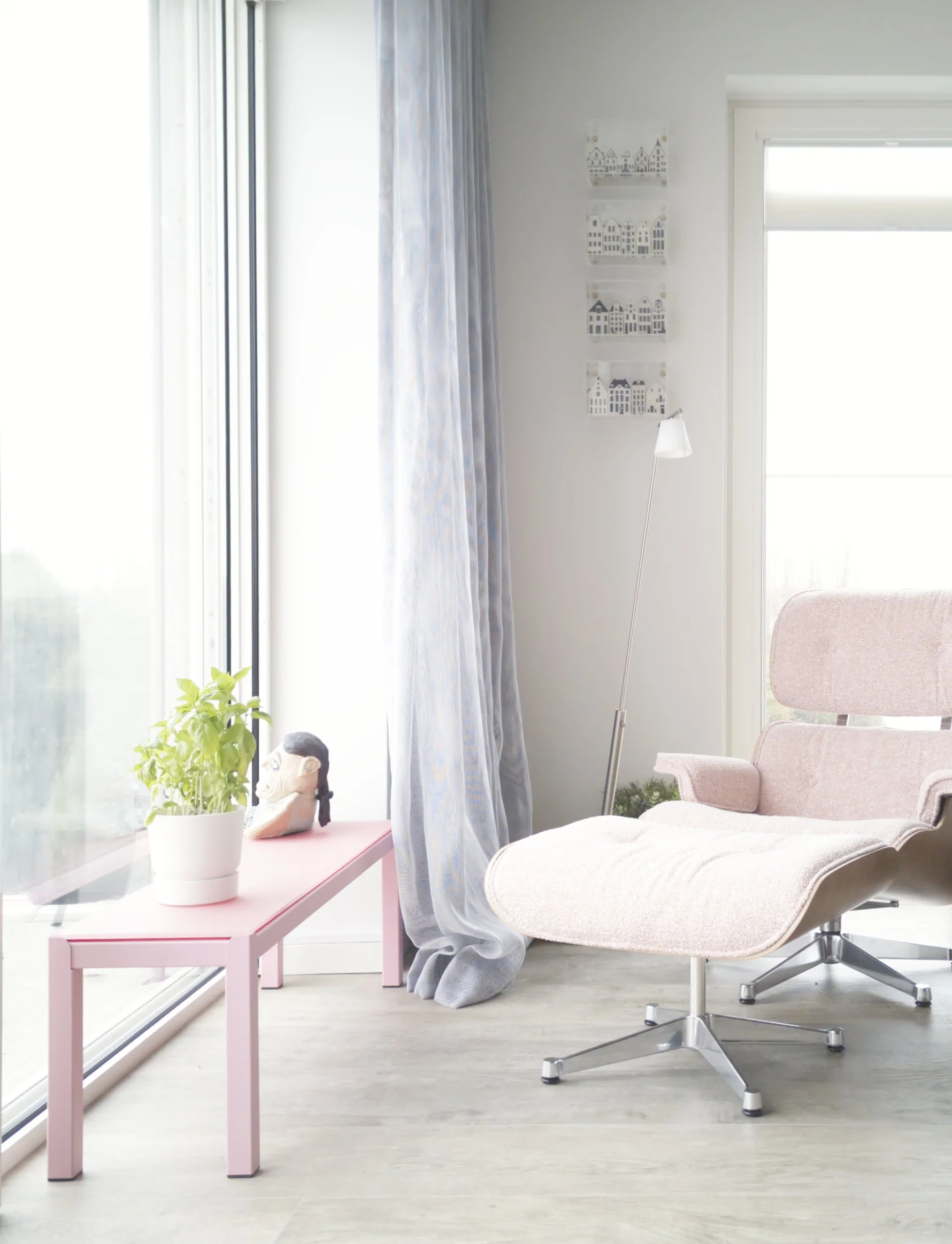 woonkamer met roze tafel, planten, zwarte dierbeeldje, lichte fauteuil, grote ramen met blauwe gordijnen, muurdecoratie