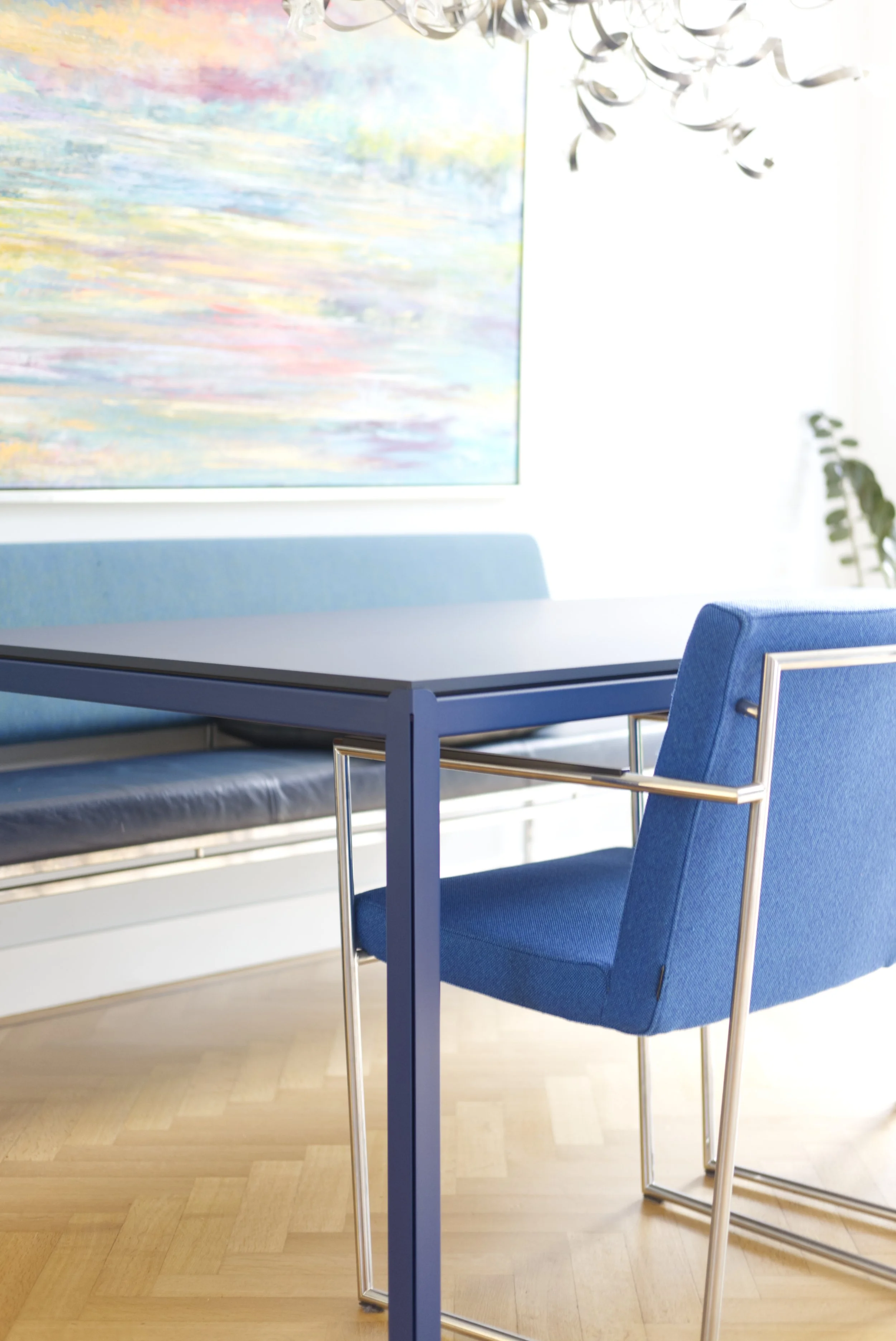 Een blauwe fauteuil met metalen armleuningen naast een zwart tafel in een moderne, lichte kamer met houten vloer, een kleurrijk abstract schilderij aan de muur en een plafondlamp.