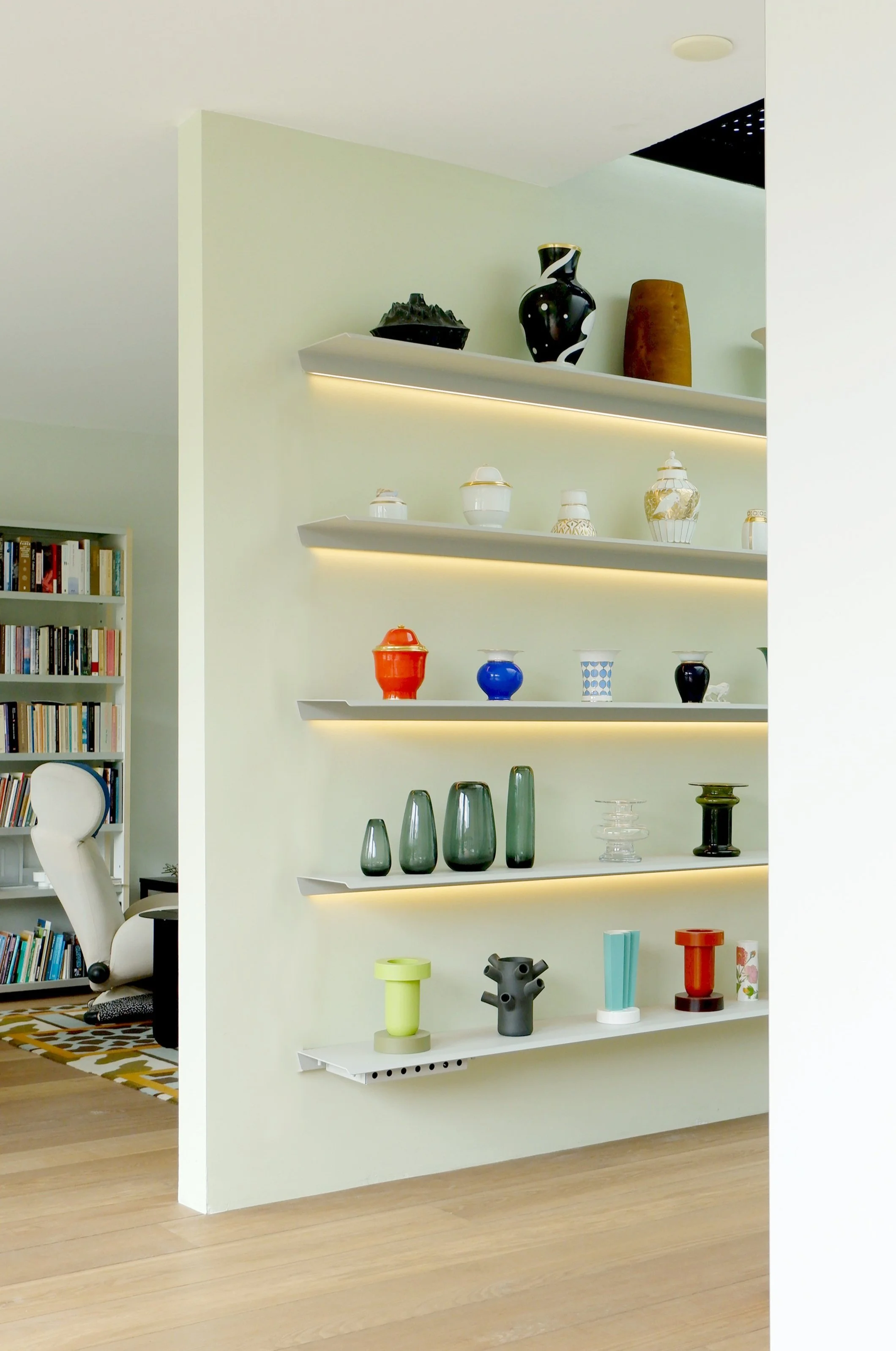 Witte open plank met ingebouwde verlichting met diverse kleurrijke vazen en decoratieve objecten in een modern interieur met houten vloer en boekenkast in de achtergrond.