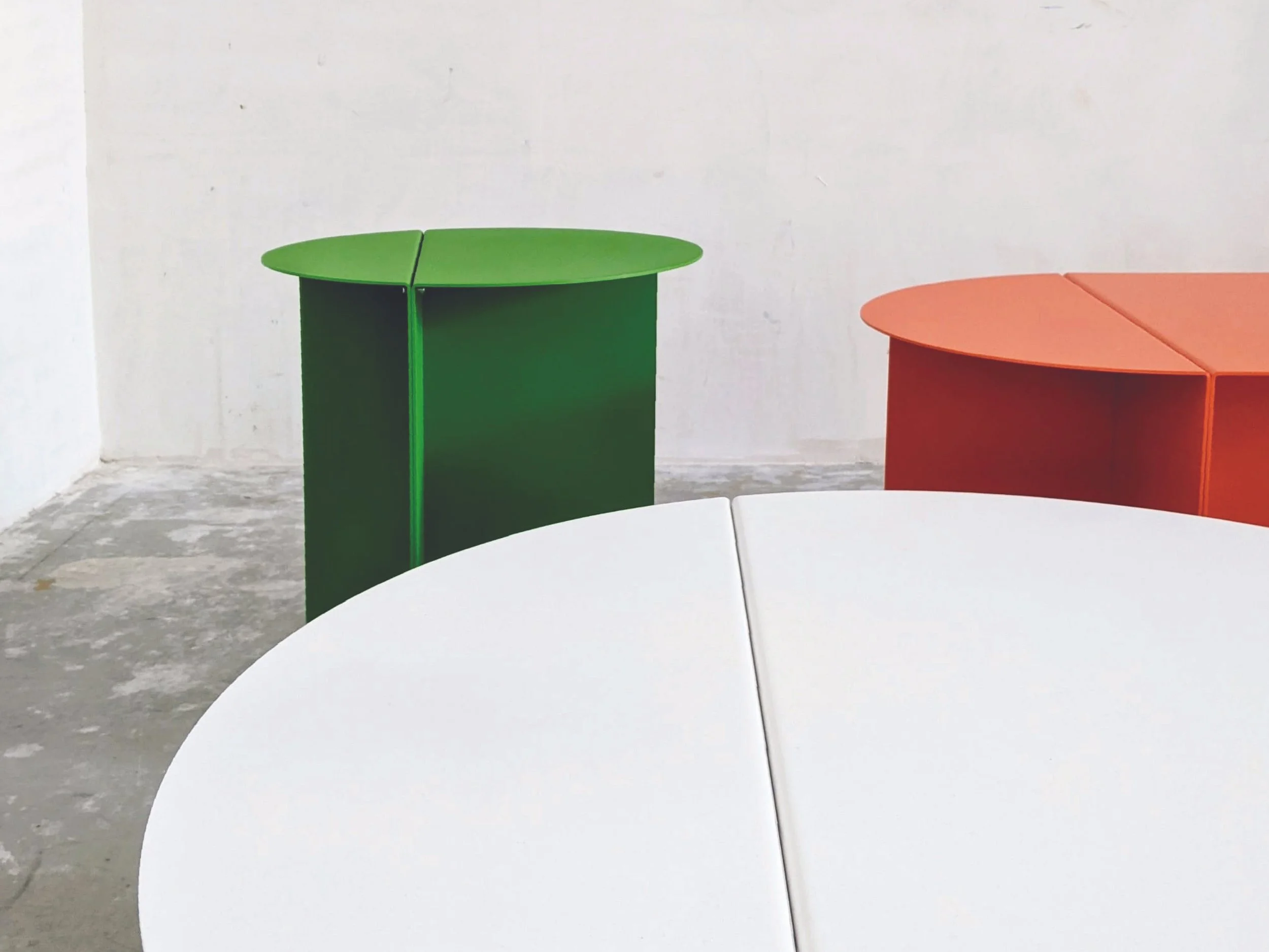 Een witte ronde tafel en twee kleurrijke aparte ronde kasten, één groen en één oranje, in een minimalistische kamer met een witte muur en een betonnen vloer.