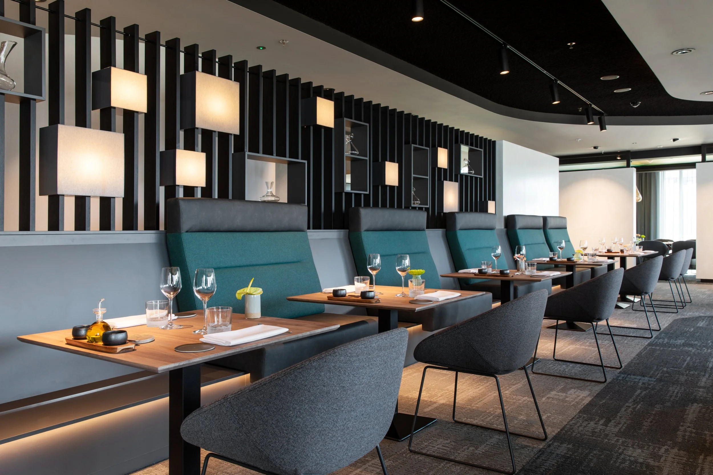Interieur van een modern restaurant met wit en zwart decor, groene stoelen, houten tafels en gedekte borden en glaswerk.