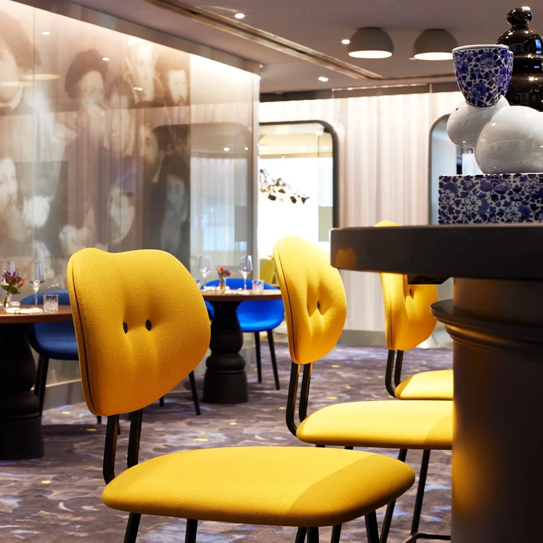 Een restaurant met gele en blauwe stoelen, een tafel met glazen en bloemen, en kleurrijke vazen en bakjes op een bar.