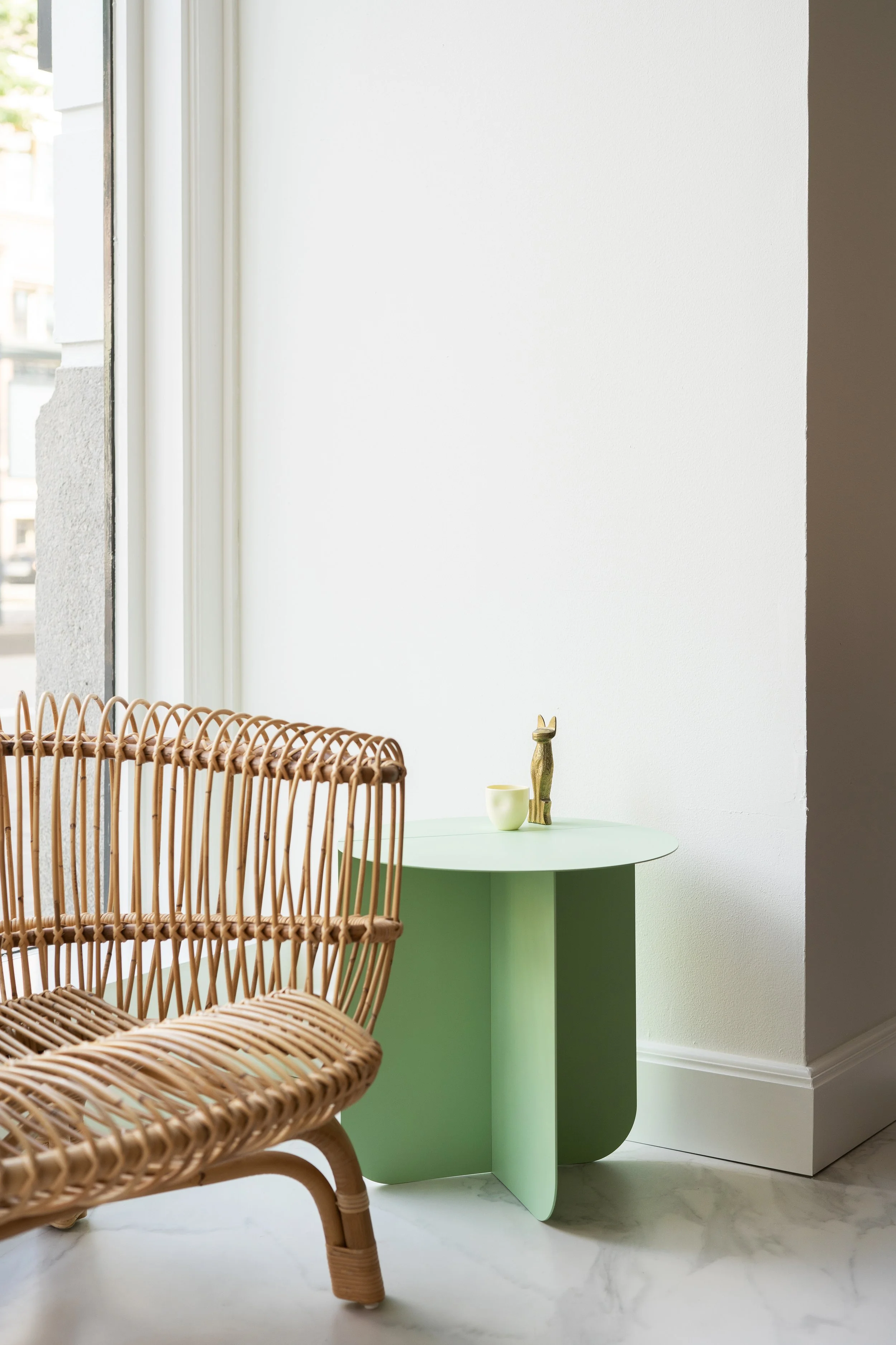 Een binnenruimte met een rieten stoel, een lichtgroene bijzettafel met decoratieve objecten, een witte muur, en een groot raam dat natuurlijk licht binnenlaat.