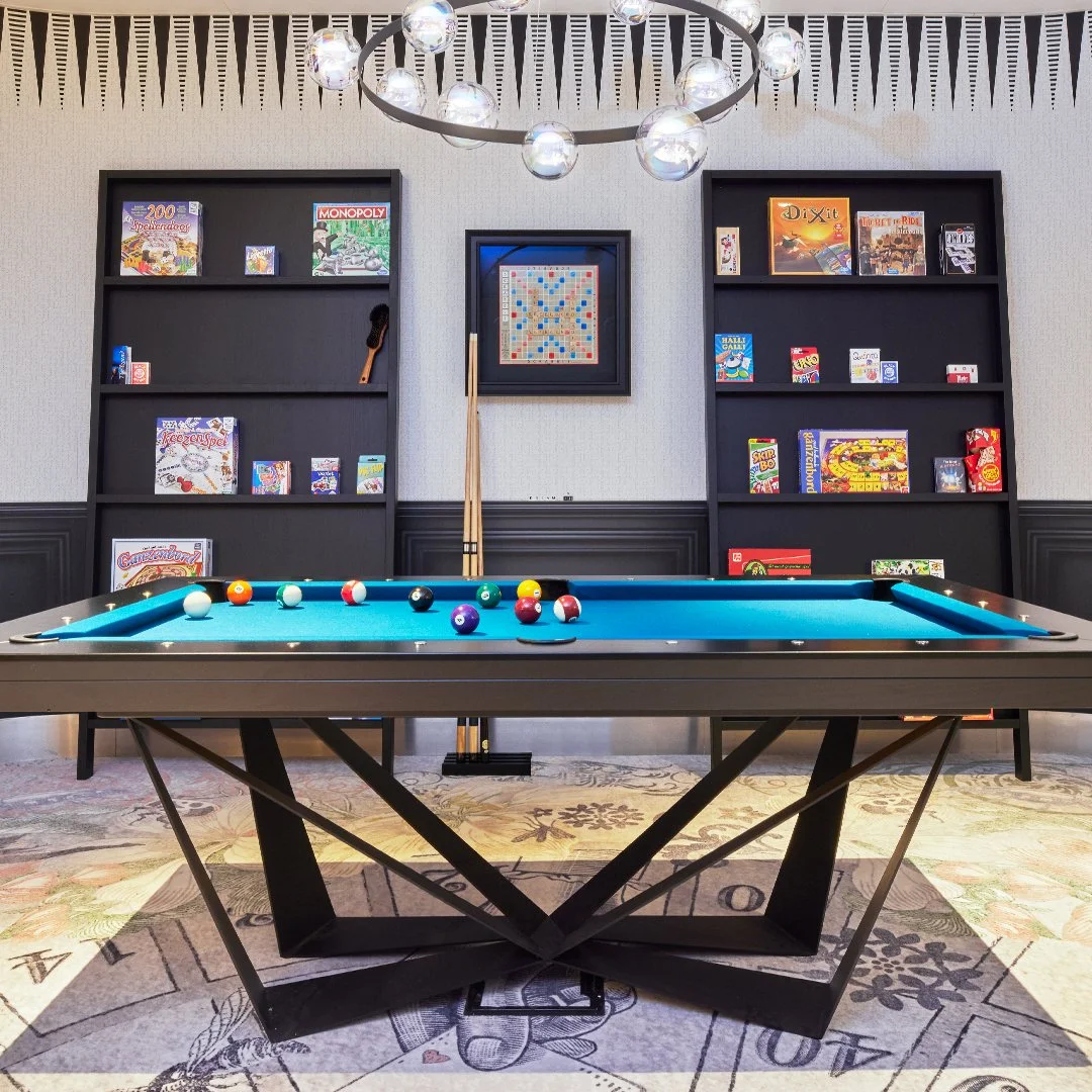 Bureau met biljarttafel, omringd door boekenkasten met bordspellen, verlichte kroonlamp, en een muur met een zwart-wit patroon.