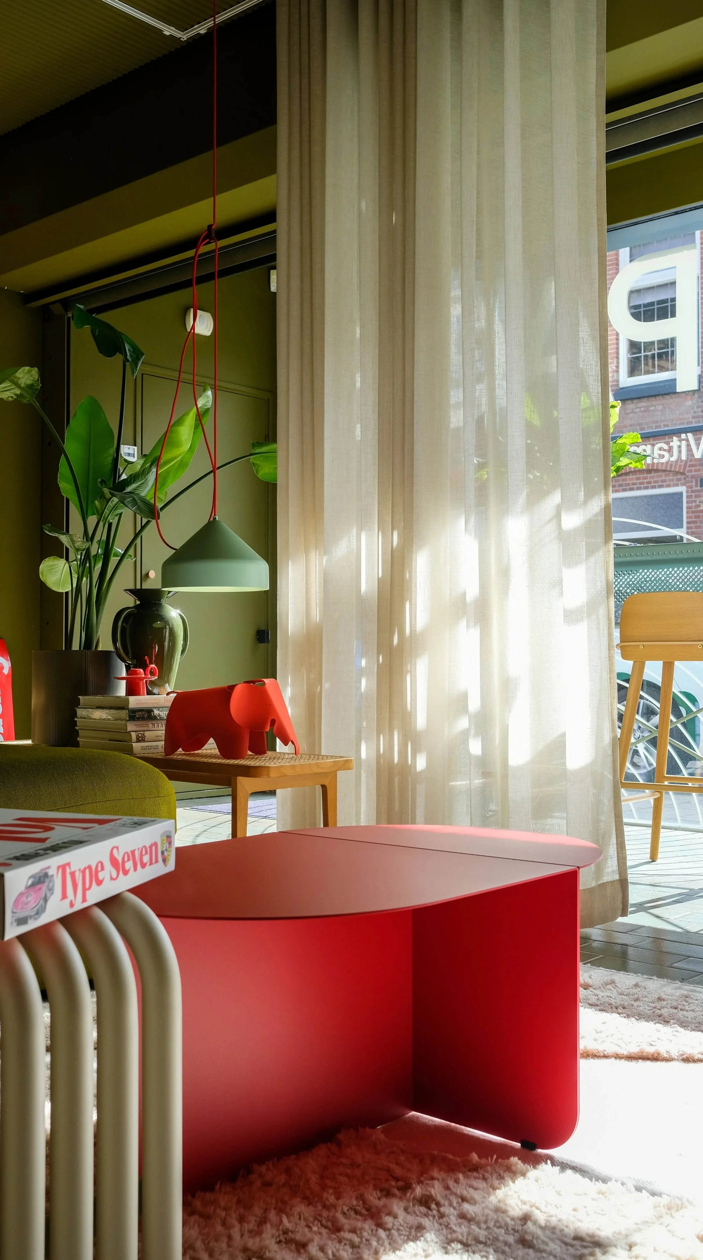 Binnenruimte met groene plant, rode en groene decoraties, een boekenkast, en een groot raam met witte gordijnen dat veel natuurlijk licht binnenlaat.