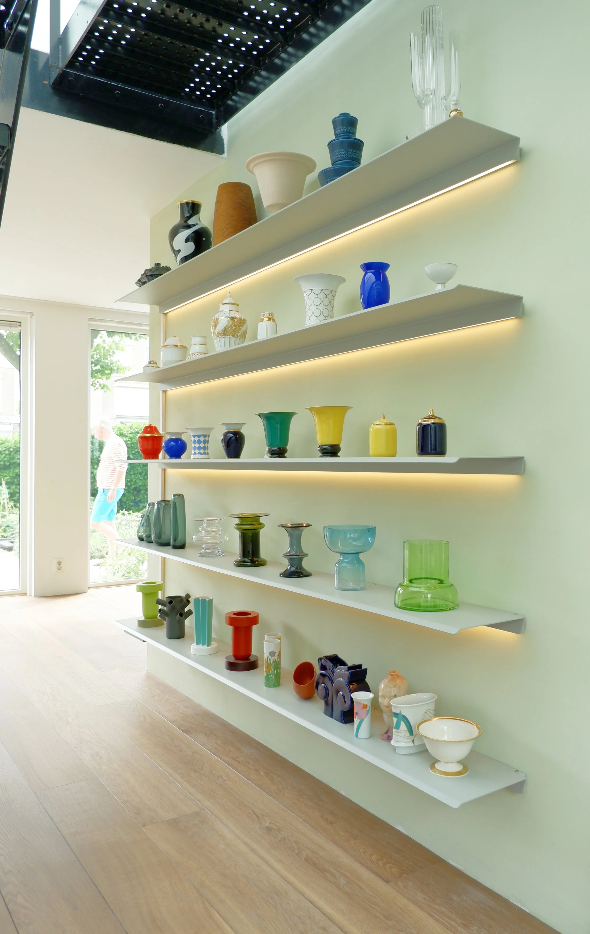 Verschillende kleurrijke glazen en vazen op witte planken tegen een lichtgroene muur, met een grote raam en een persoon zichtbaar buiten.