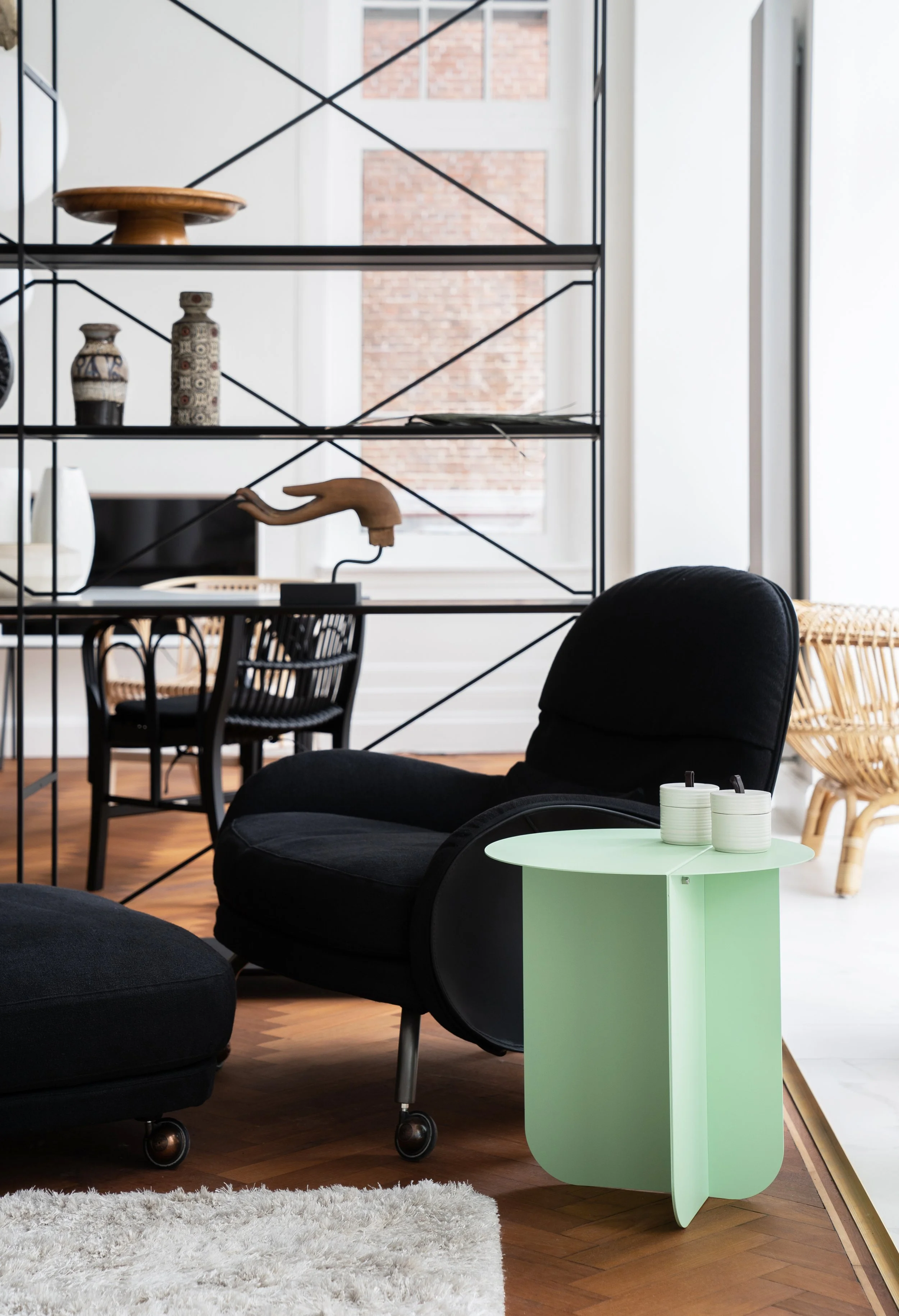 Een moderne woonkamer met een zwart fauteuil en een groene bijzettafel met twee potjes, een glazen wand en een zwarte metalen boekenplank met decoratieve vazen en een houten handmodel, in een lichte ruimte met grote ramen.