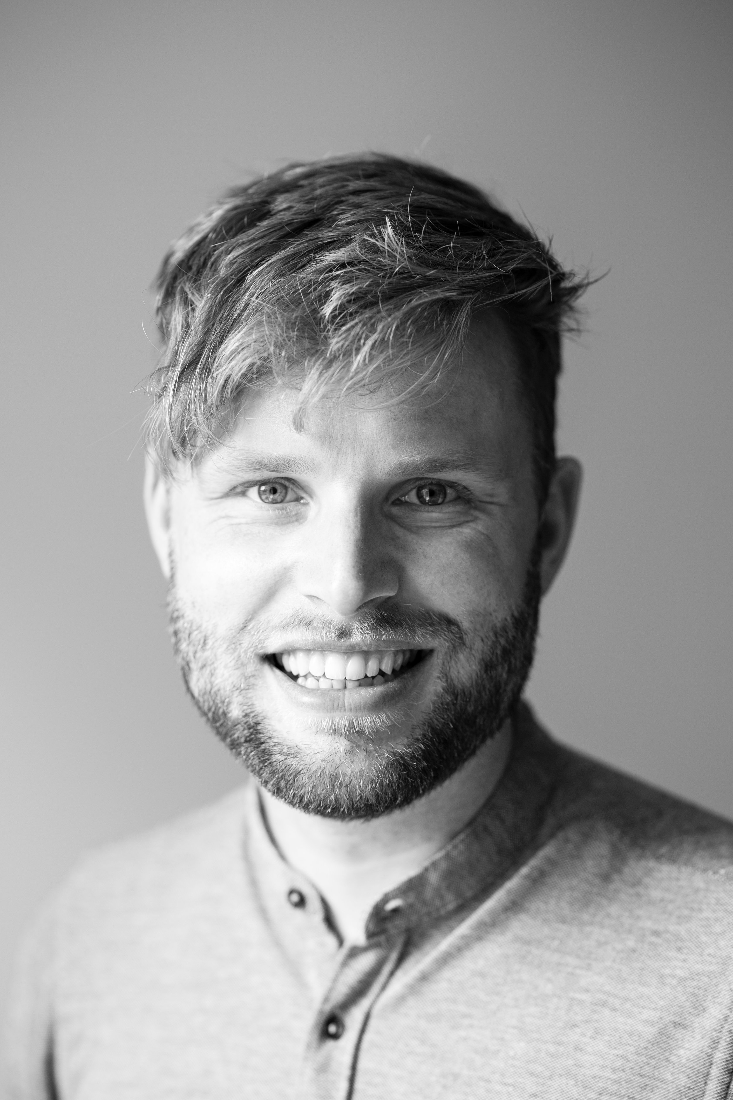 Zwart-wit portret van een lachende man met korte haar en baard, gekleed in een colbert met knoopsluiting