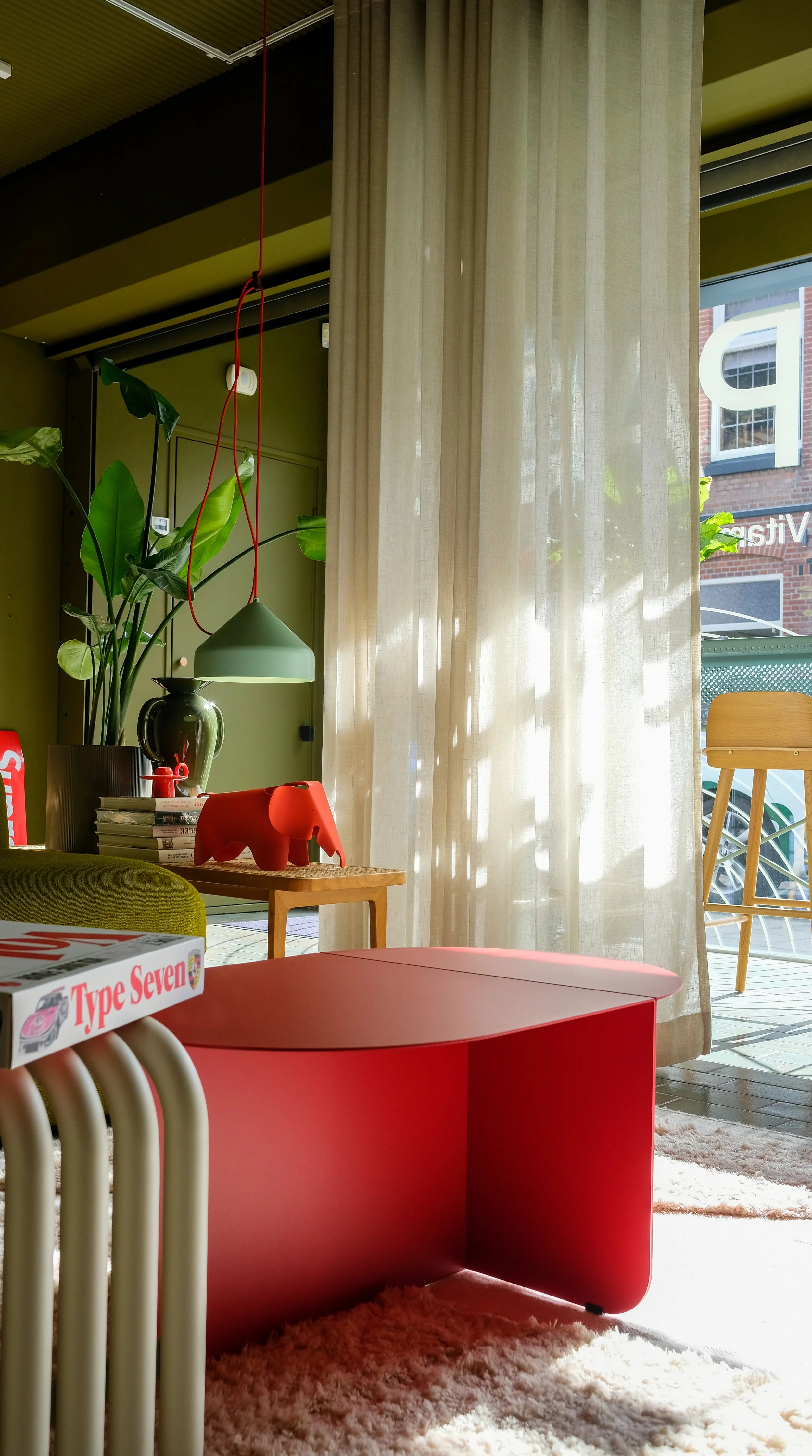 Gezellige interieurruimte met een grote plant, een groene hanglamp, een rood diermodel, een paar boeken, een grote groene vaas, een rood bijzettafeltje, en een grote raam met lichte gordijnen die zonlicht binnenlaten.