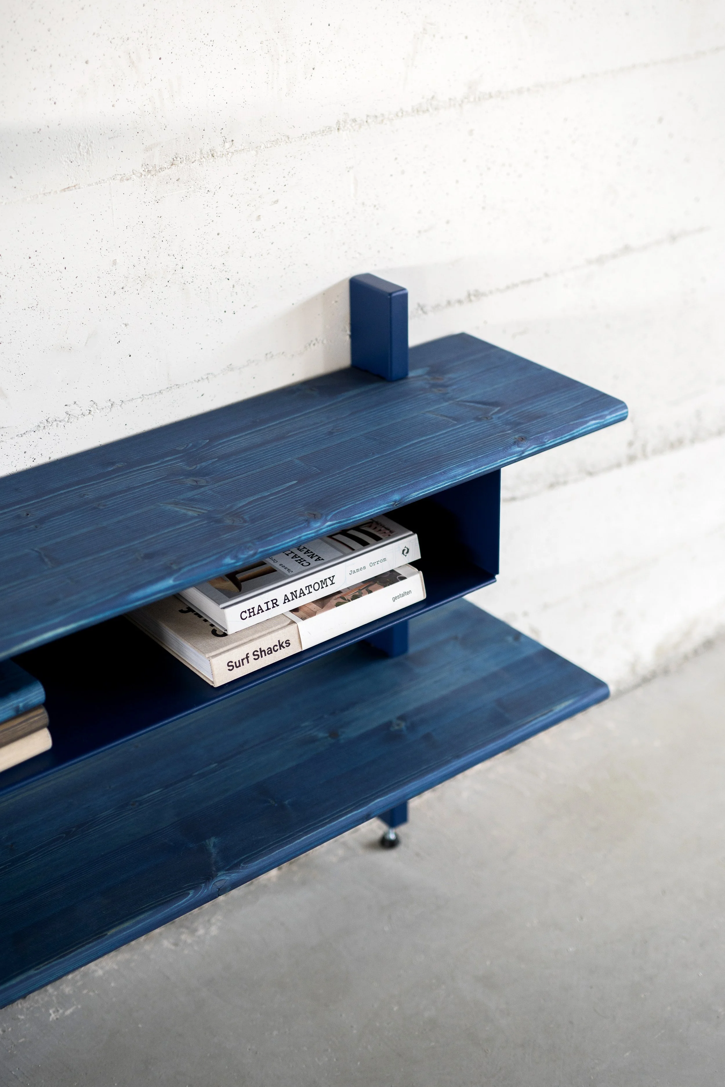 Een blauwe wandbank met boeken, waaronder 'Chair Anatomy' en 'Surf Shacks', tegen een witte muur met een betonnen vloer.