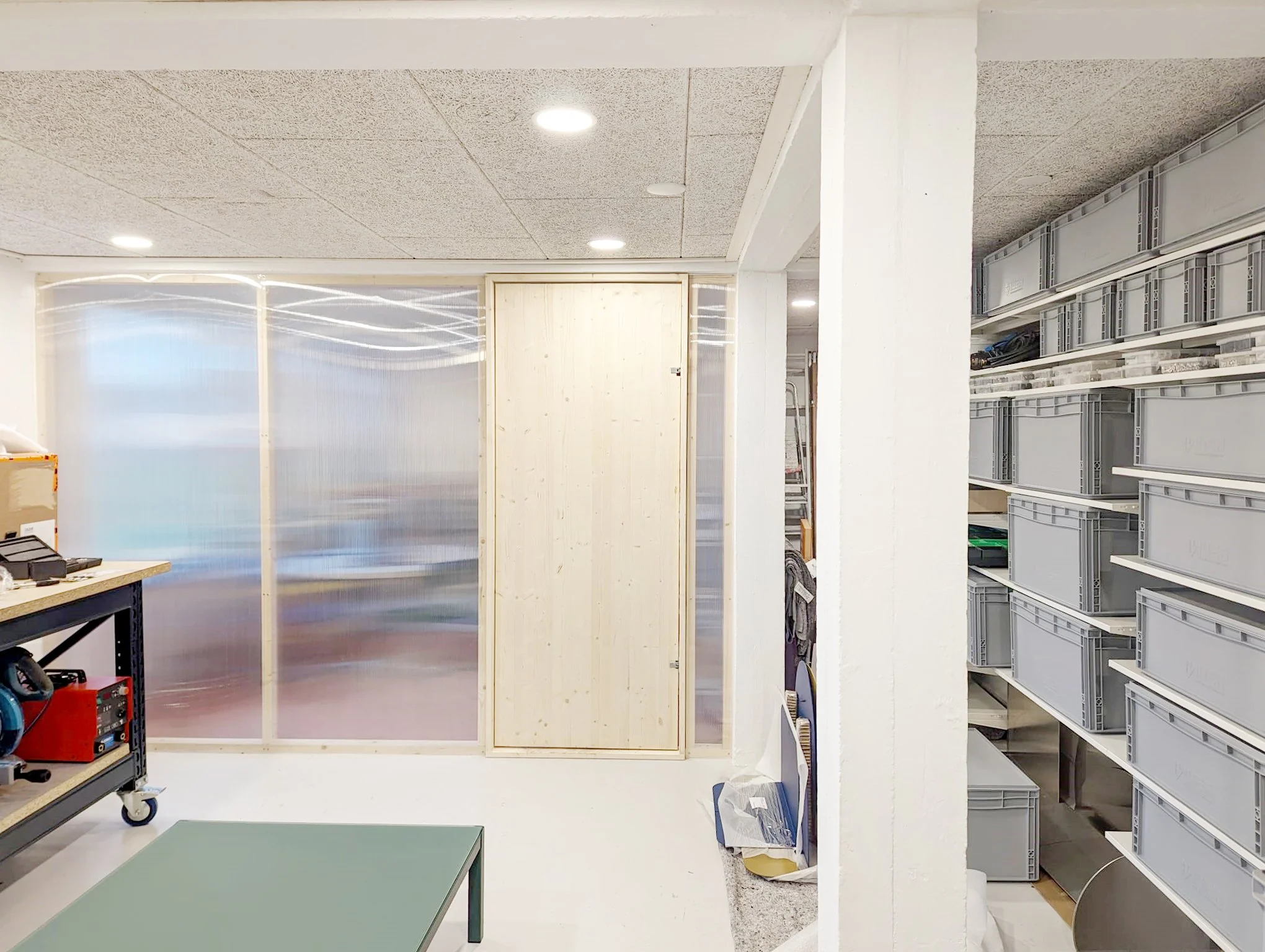 Binnenruimte met metalen opslagrekken, een werktafel en een houten deur, verlicht door plafondverlichting.